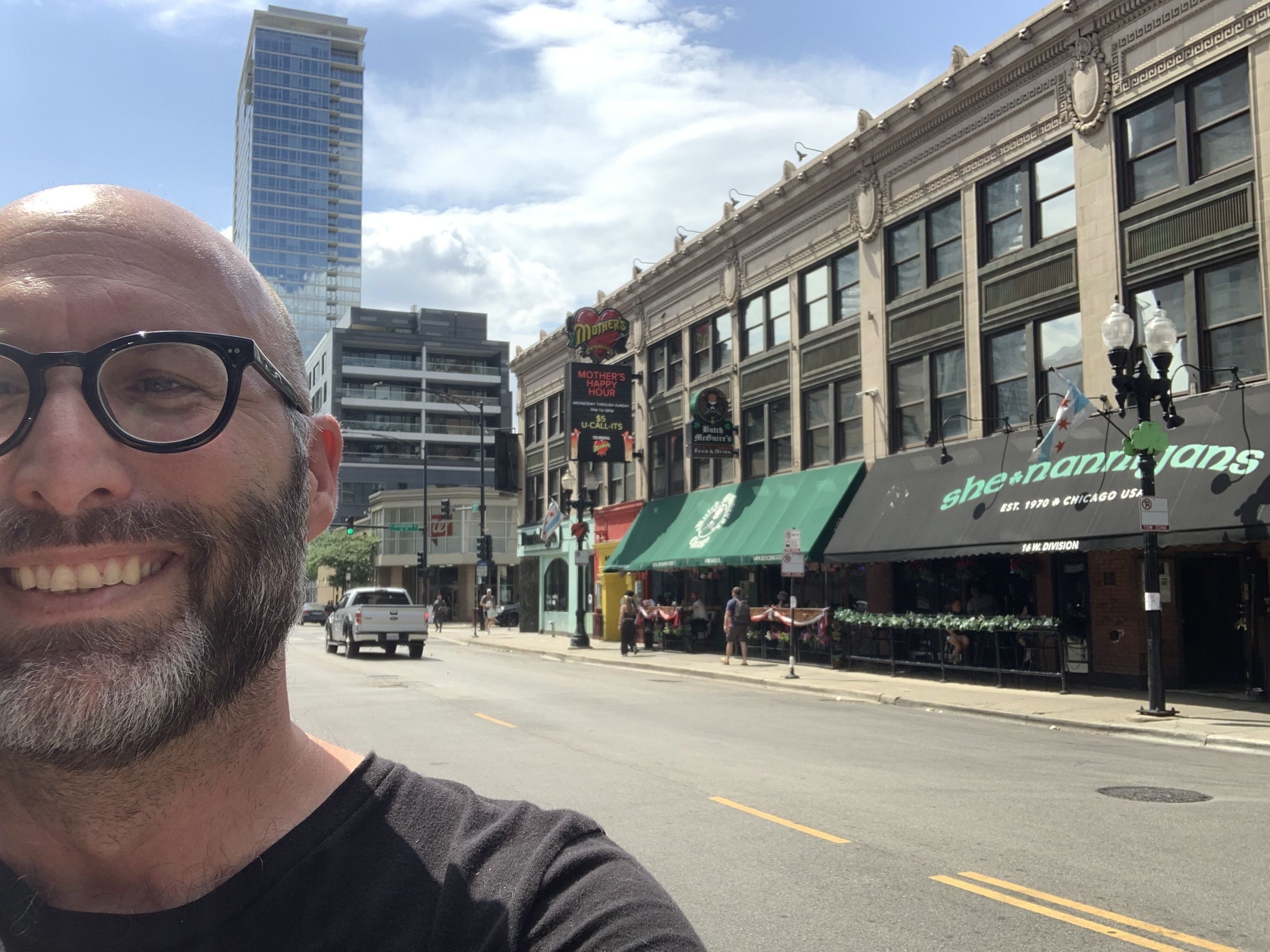 Up close image of man with black glasses and beard standing across the street from a strip of bars.