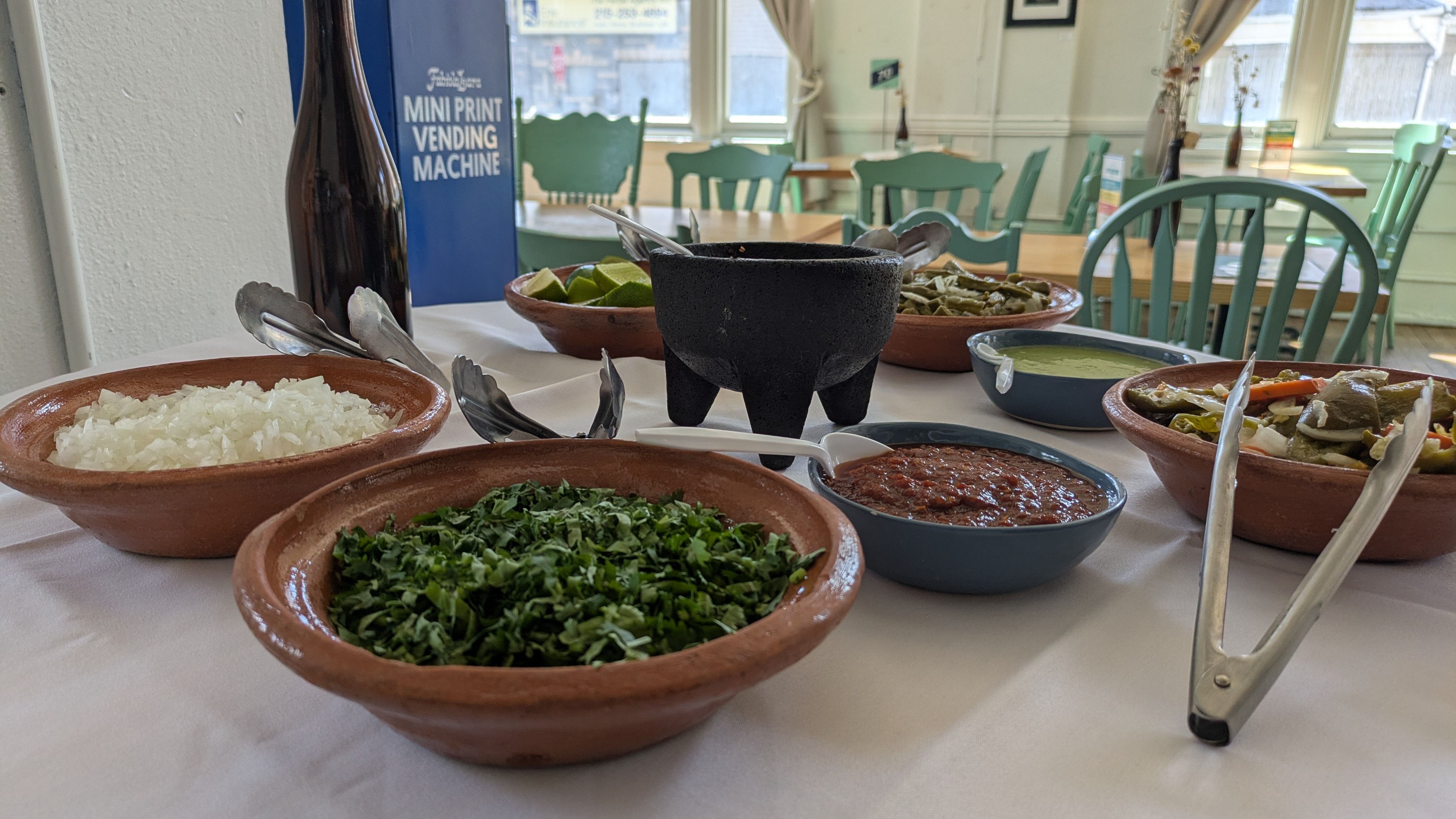 Table set with clay bowls of rice, greens, sauces and lime wedges; a central black molcajete. In the background, a blue vending machine labeled "MINI PRINT VENDING MACHINE" and teal cafe chairs.