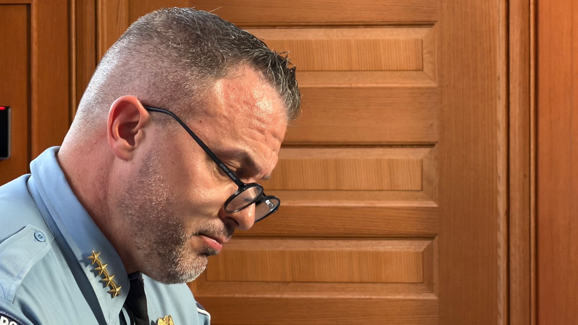 Side profile of a police officer in a light blue uniform with a gold badge and black glasses, standing before tall wooden paneled doors.