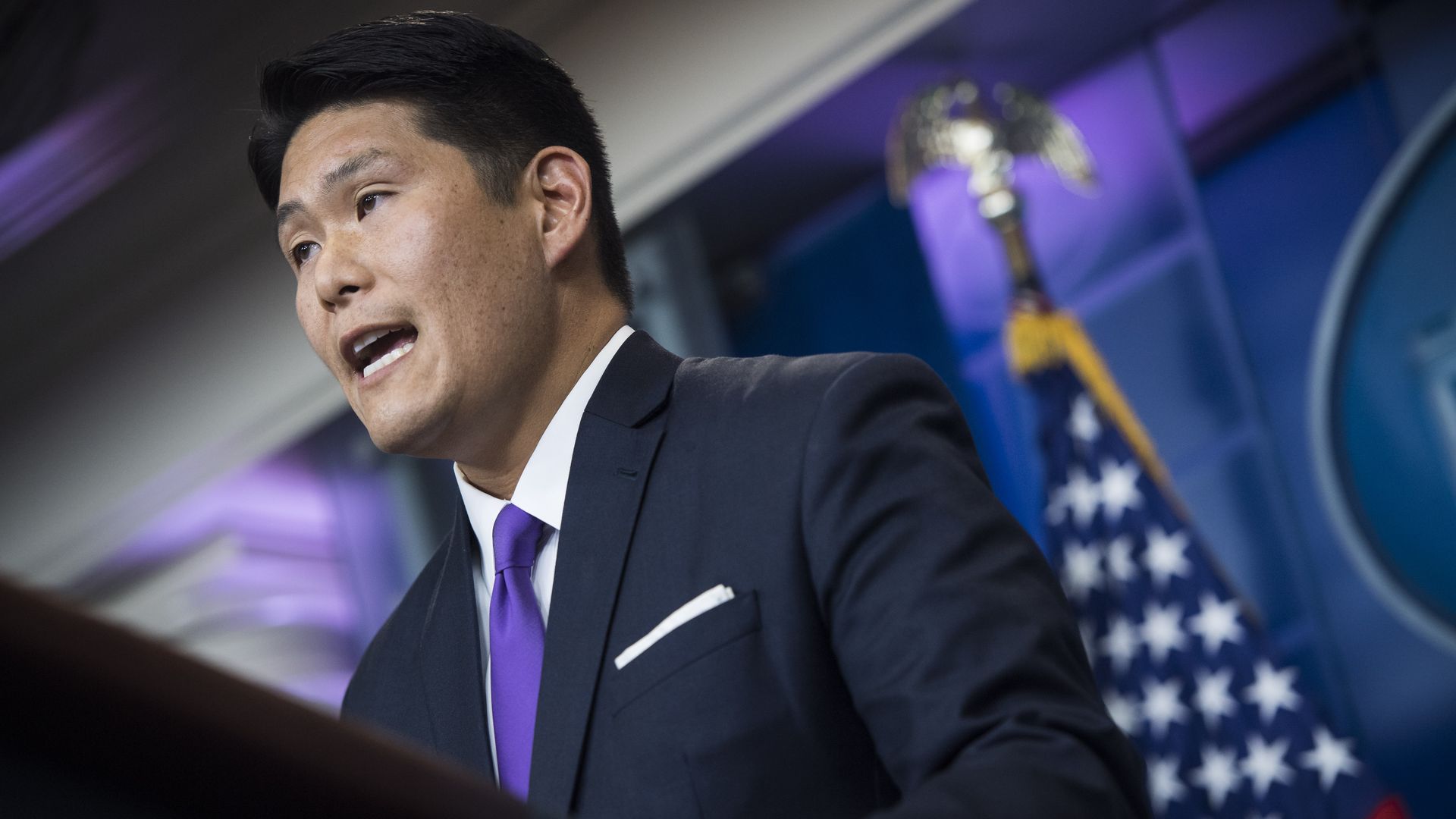 Robert Hur, wearing a dark blue suit jacket, white shirt and purple tie, stands in front of a blue backdrop and an American flag in the White House press briefing room.