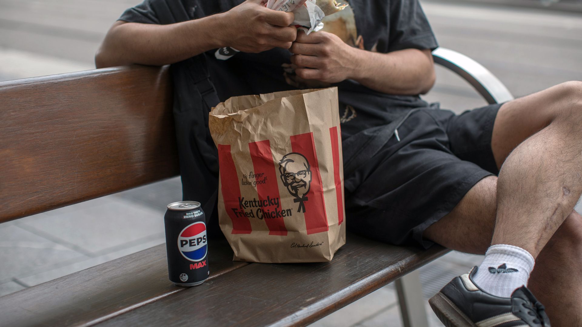 A man in a dark T-shirt sits on a wooden bench, unwrapping a sandwich. A KFC paper bag and a Pepsi Max can are on the bench beside him in an urban street setting.