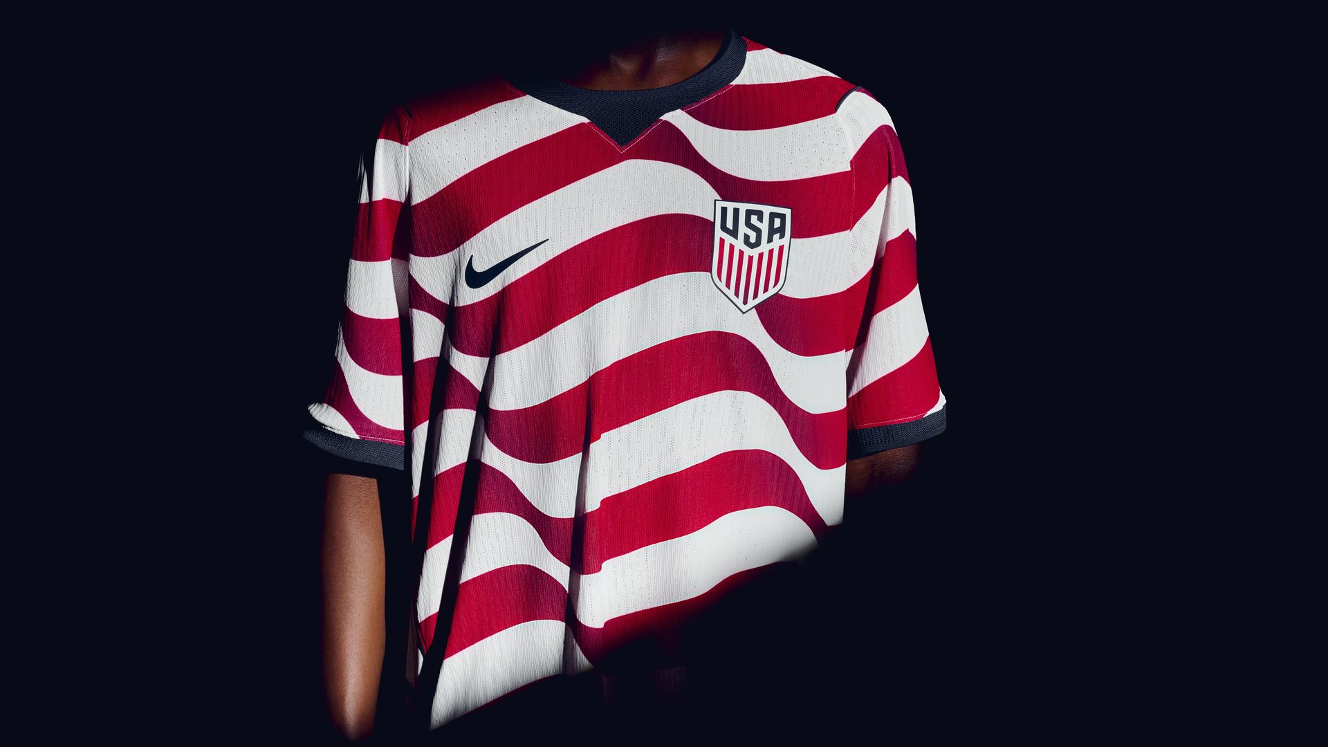 Person wearing a red-and-white wavy striped USA soccer jersey with a navy collar, a dark Nike swoosh, and the USA shield crest, set against a black background.