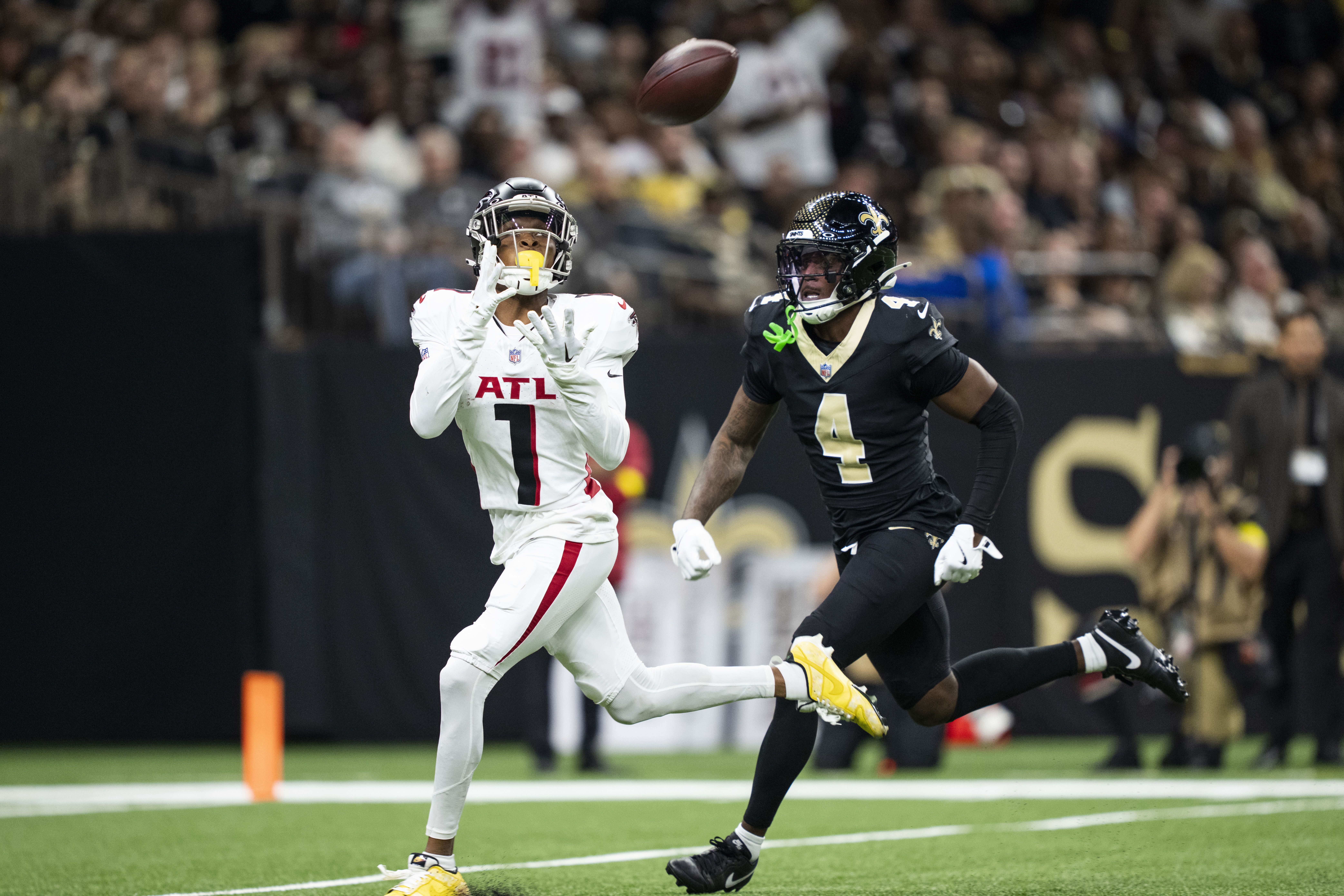 Two NFL players in action: one in white Atlanta Falcons uniform catching a football, and one in black New Orleans Saints uniform running behind him on a football field.