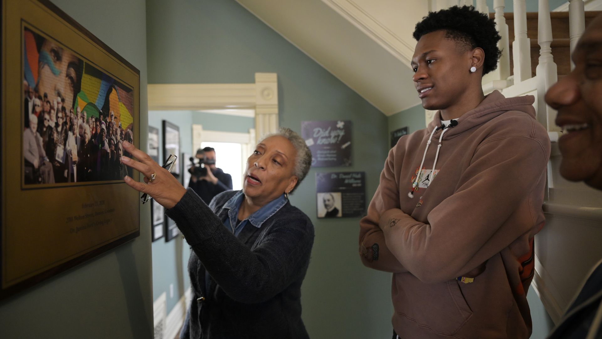 A woman in a sweater points toward a portrait while standing next to a man in a hooded sweater. 