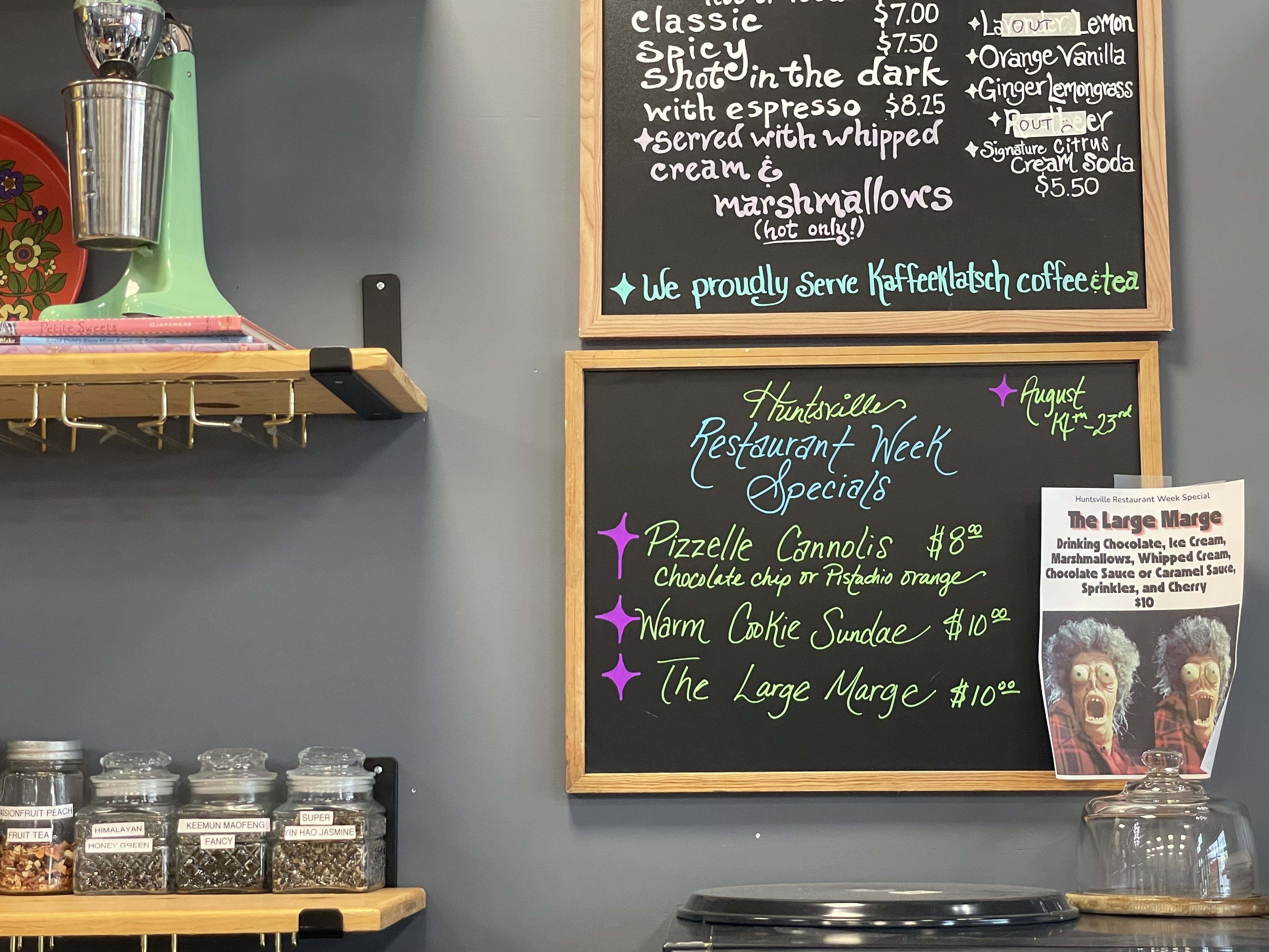 Cafe interior with a mint-green vintage mixer on a gray wall, jars on a wooden shelf, and two framed chalkboards with colorful handwritten menu items like classic spicy shot in the dark with espresso and marshmallows.