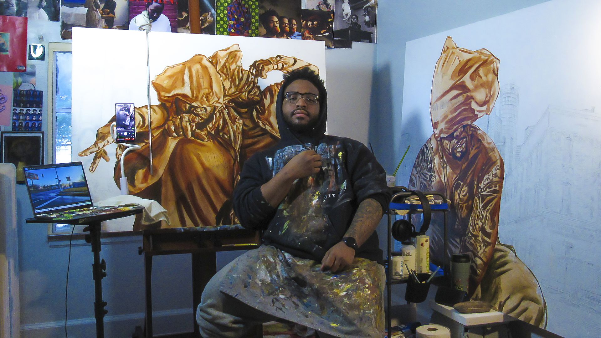 An artist wearing a paint-stained apron and hoodie sits in a studio surrounded by paintings, art and supplies. 