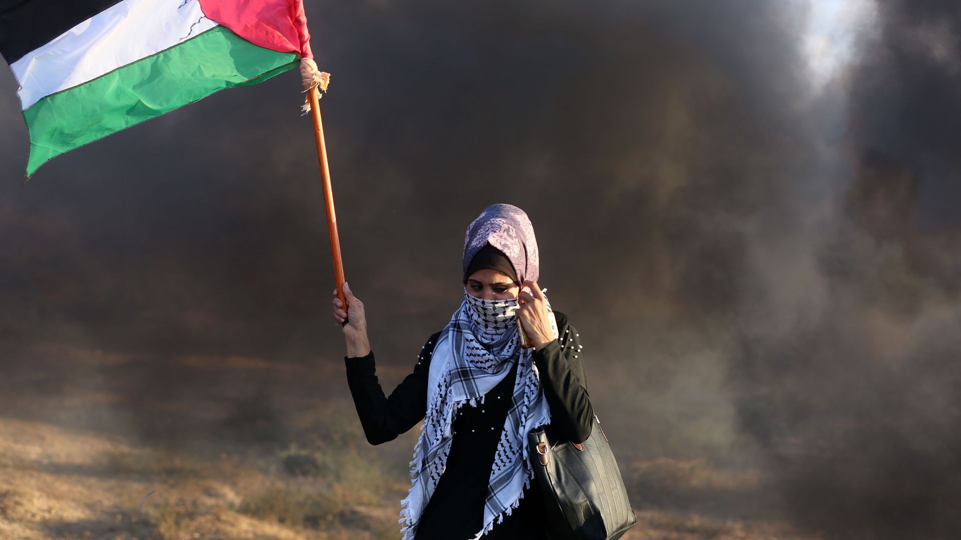 A woman holding a Palestinian flag, standing in black smoke. 