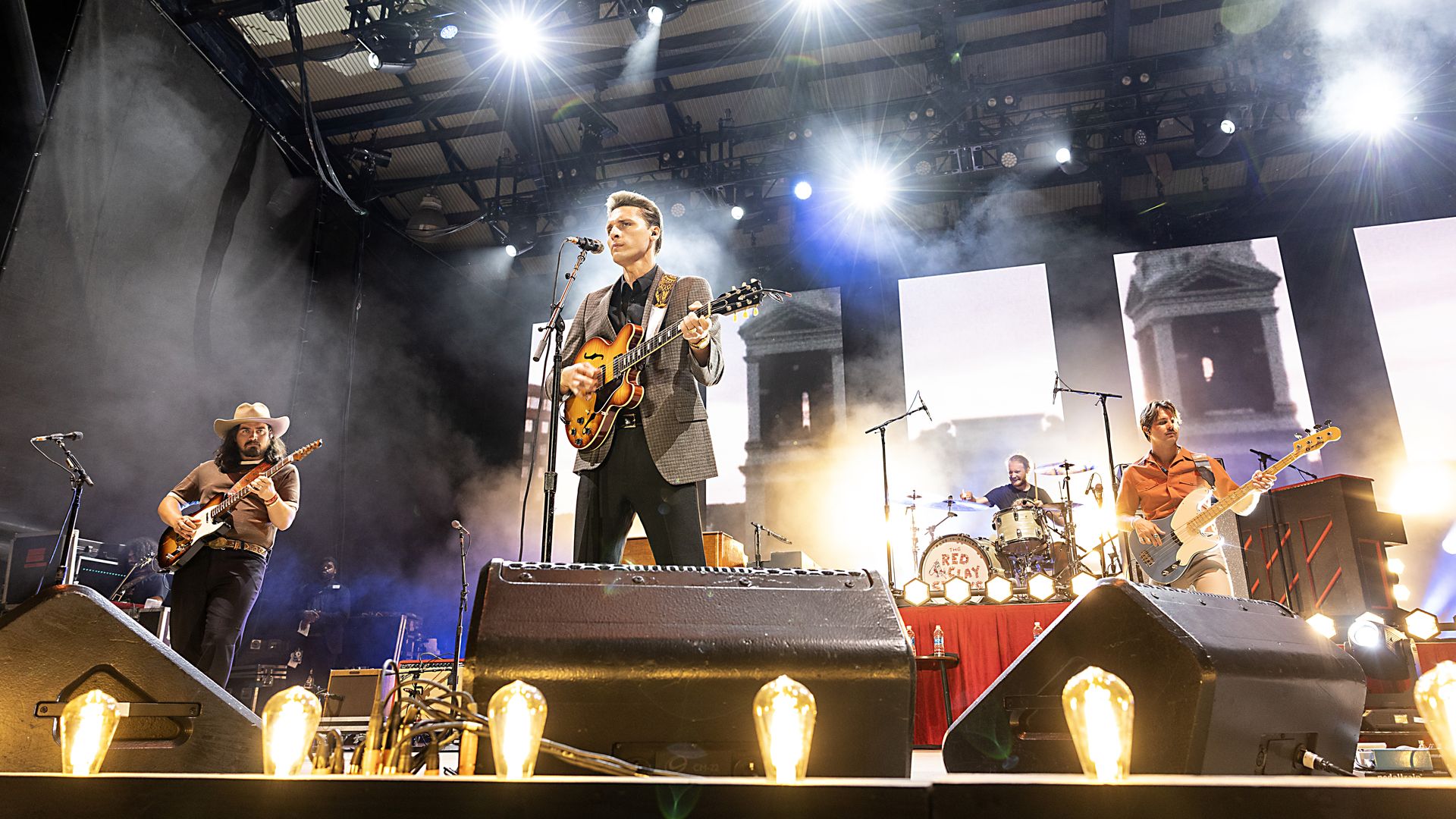 Lead singer in a gray blazer plays an orange electric guitar at center stage, flanked by fellow guitarists. Bright stage lights, smoke, and large rigging loom overhead.