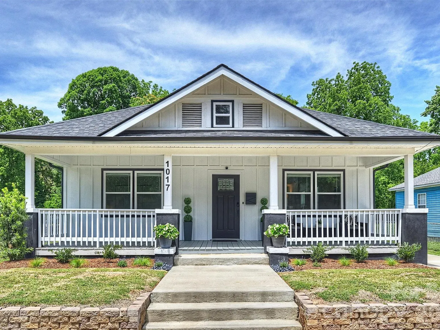 White, single-story home with a covered front porch, black door, and two large windows. Potted plants flank the entrance; gray roof, blue sky, and green trees behind.
