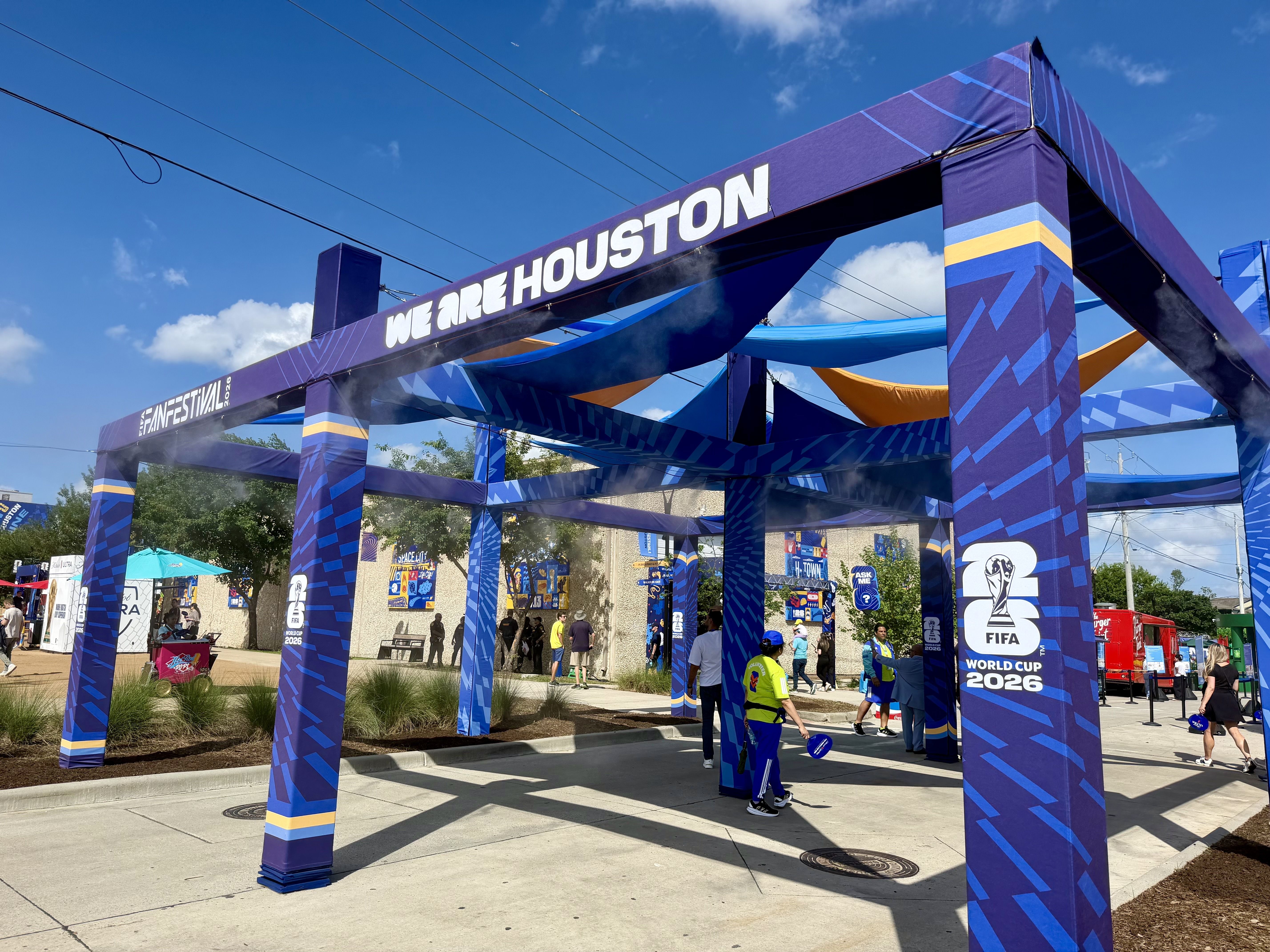 Outdoor festival entrance with a large purple arch reading "WE ARE HOUSTON" in white letters, blue and yellow decorations, FIFA World Cup 2026 signage, and people walking beneath a clear blue sky.