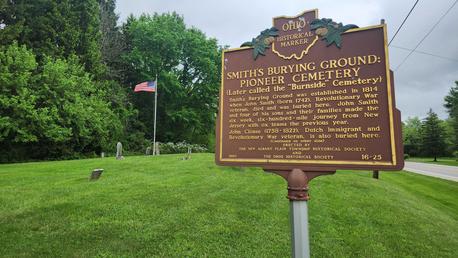 A historical marker for "Smith's Burying Ground: Pioneer Cemetery" with a waving flag and gravestones in the background.