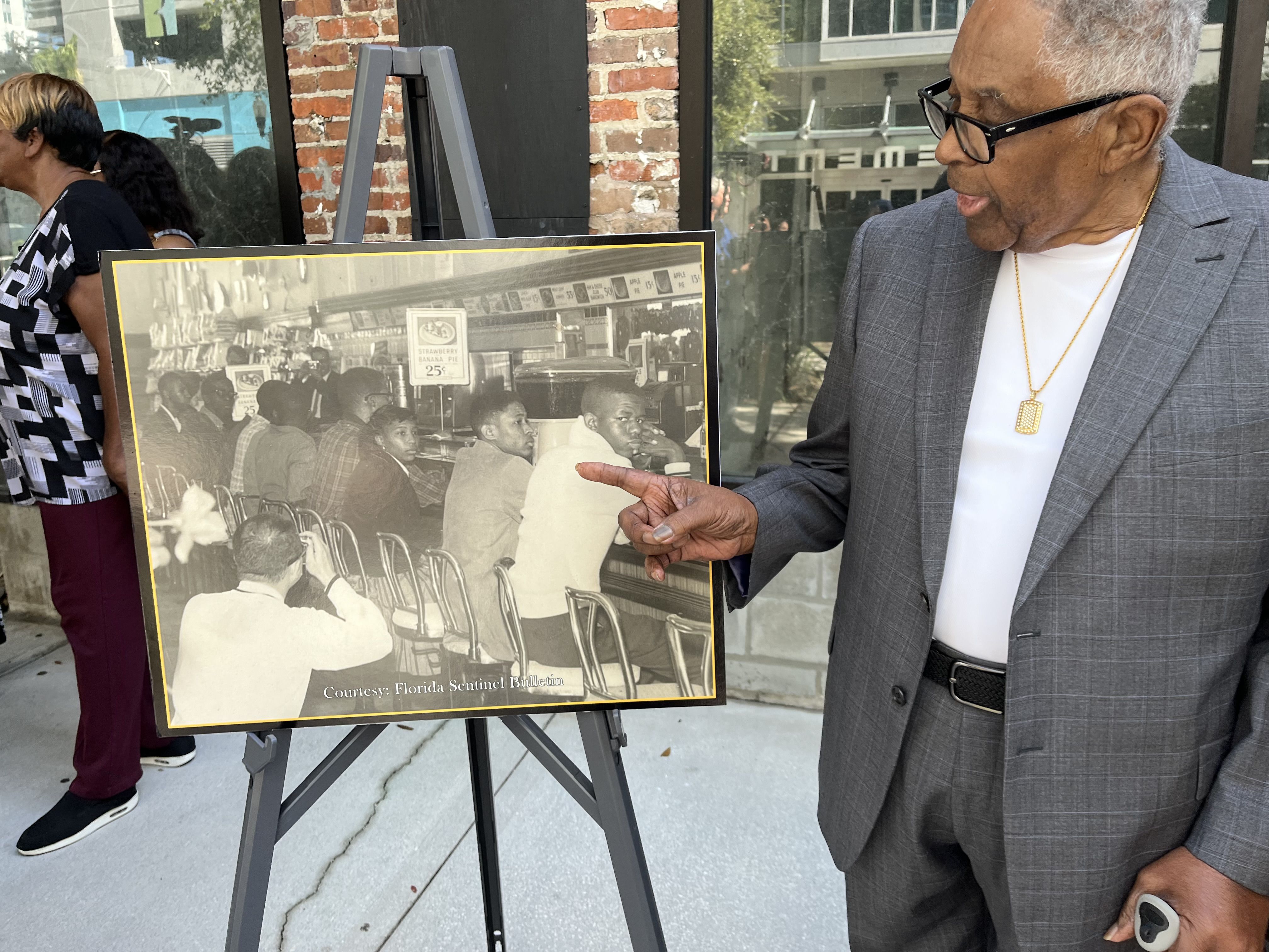 A man in a gray blazer and white T-shirt points to a boy sitting at a lunch counter in a sepia-toned photograph.