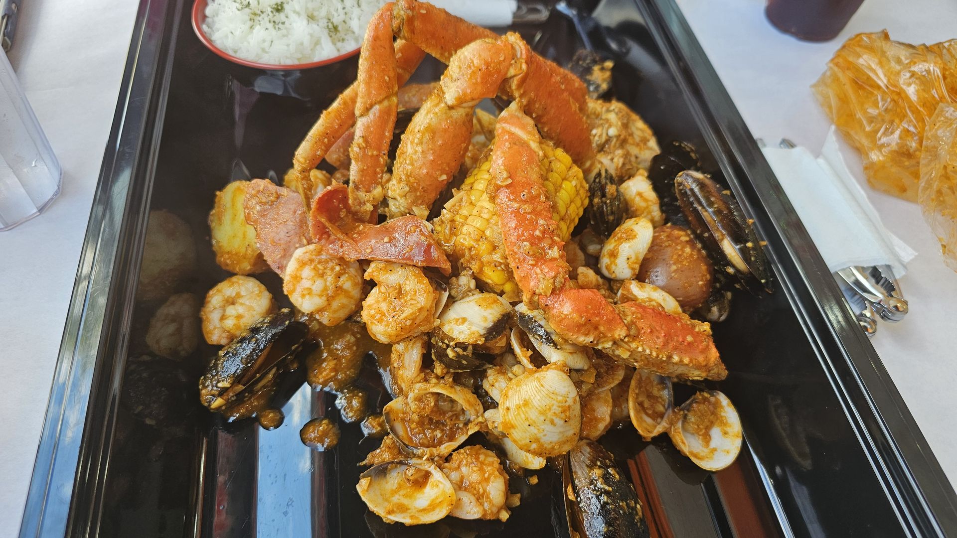 Tray with seafood boil including crab legs, shrimp, clams, mussels, corn on the cob, sausage, potatoes, and a bowl of white rice on the side.