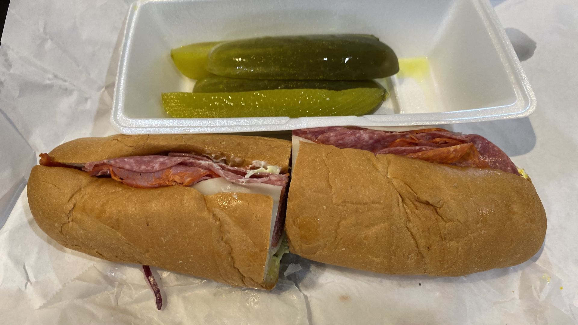 A sub sandwich cut in half next to a styrofoam container with four pickle spears.