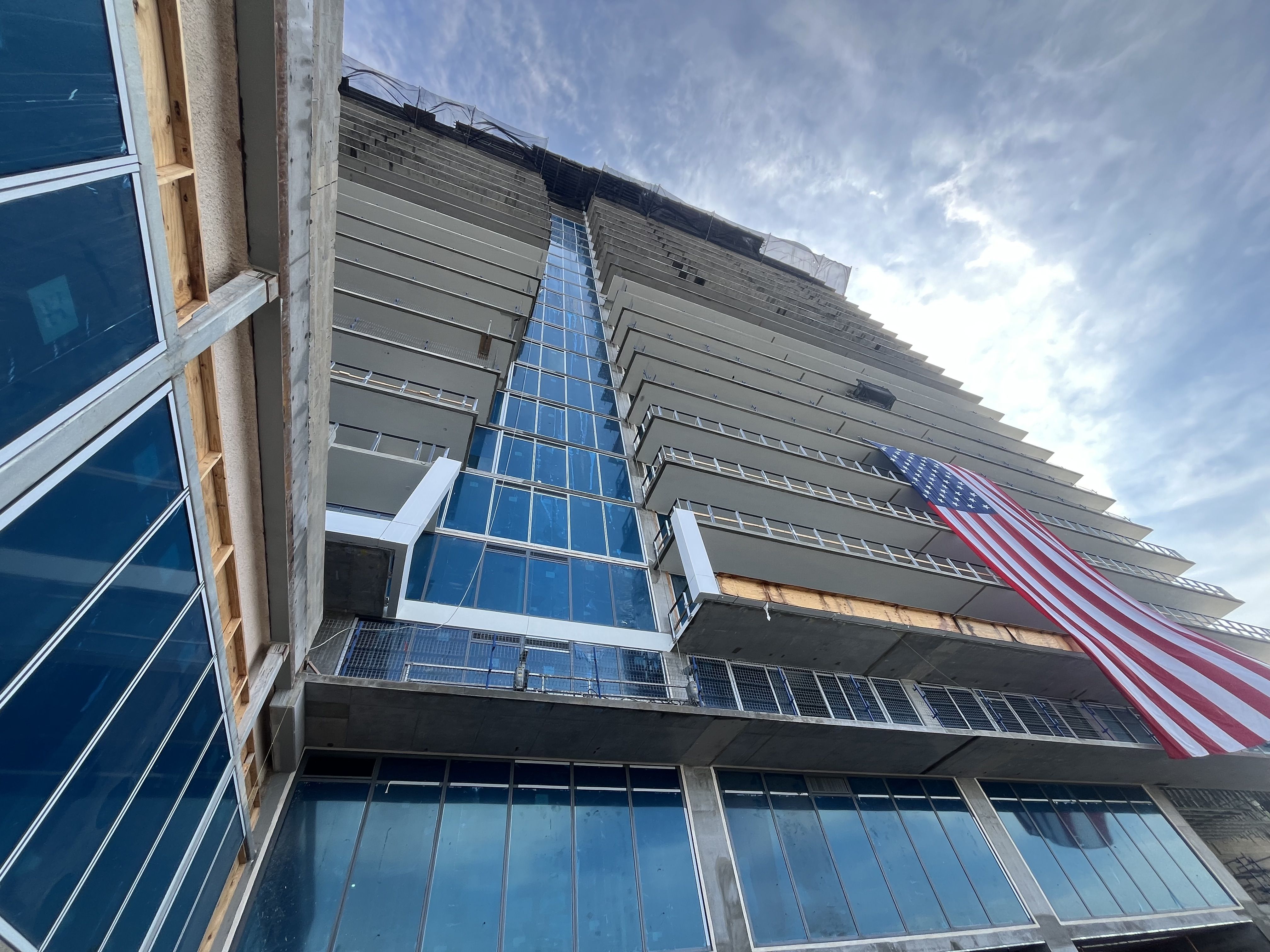 Tall modern building under construction with blue-tinted glass windows and gray concrete panels. Exposed wood framing on the left, metal railings, and an American flag draped from a balcony against a partly cloudy sky.