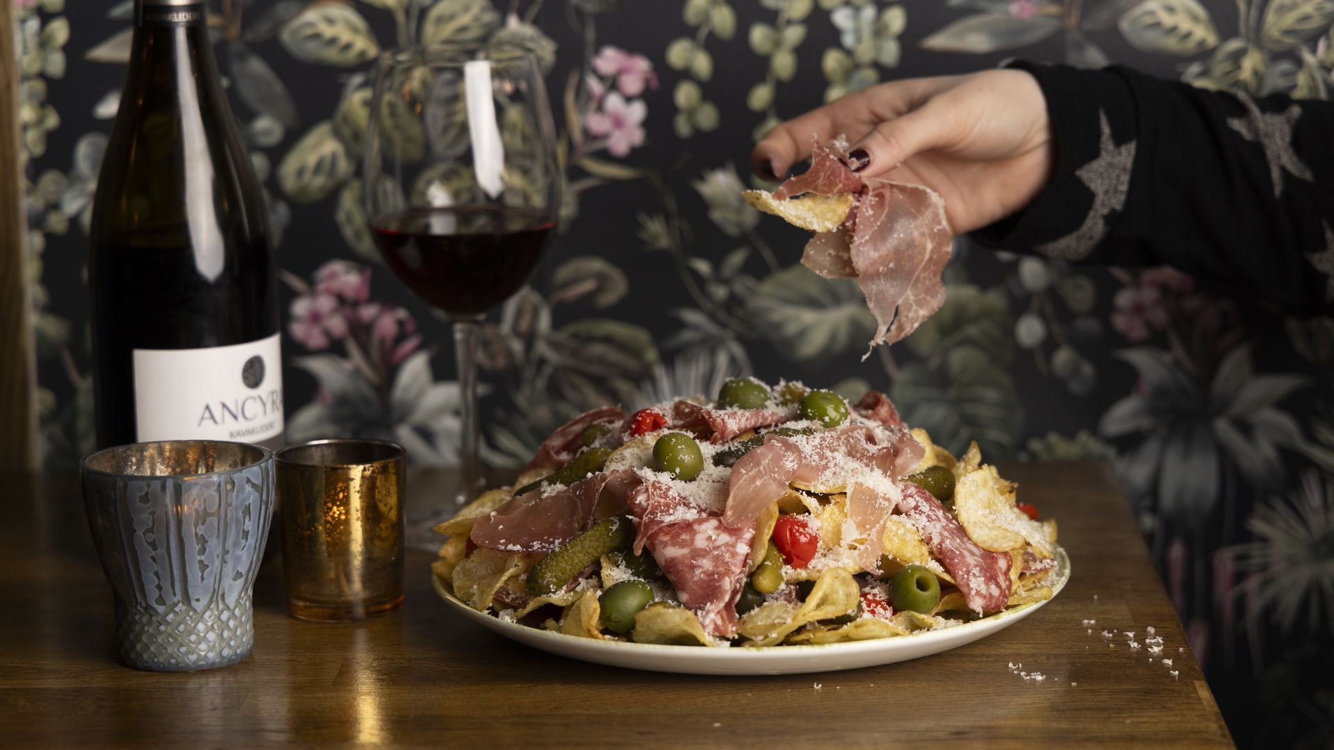 Plate of potato chips topped with sliced salami, green olives, pickles, and grated cheese on a wooden table with a bottle of red wine, filled glass, and two candles; hand holding a chip.