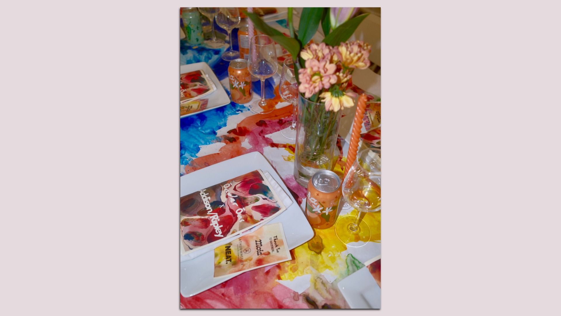 Colorful party table scene with a watercolor-pattern tablecloth, pink bouquet in a tall vase, wine glasses, an orange can, magazines, and a tall orange candle.
