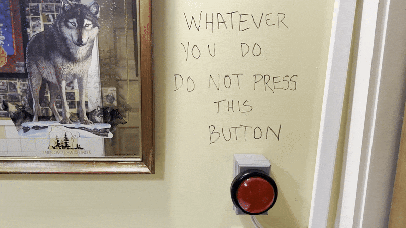 A hand reaches in and presses a button underneath a handwritten sign that says "Whatever you do do not press this button."