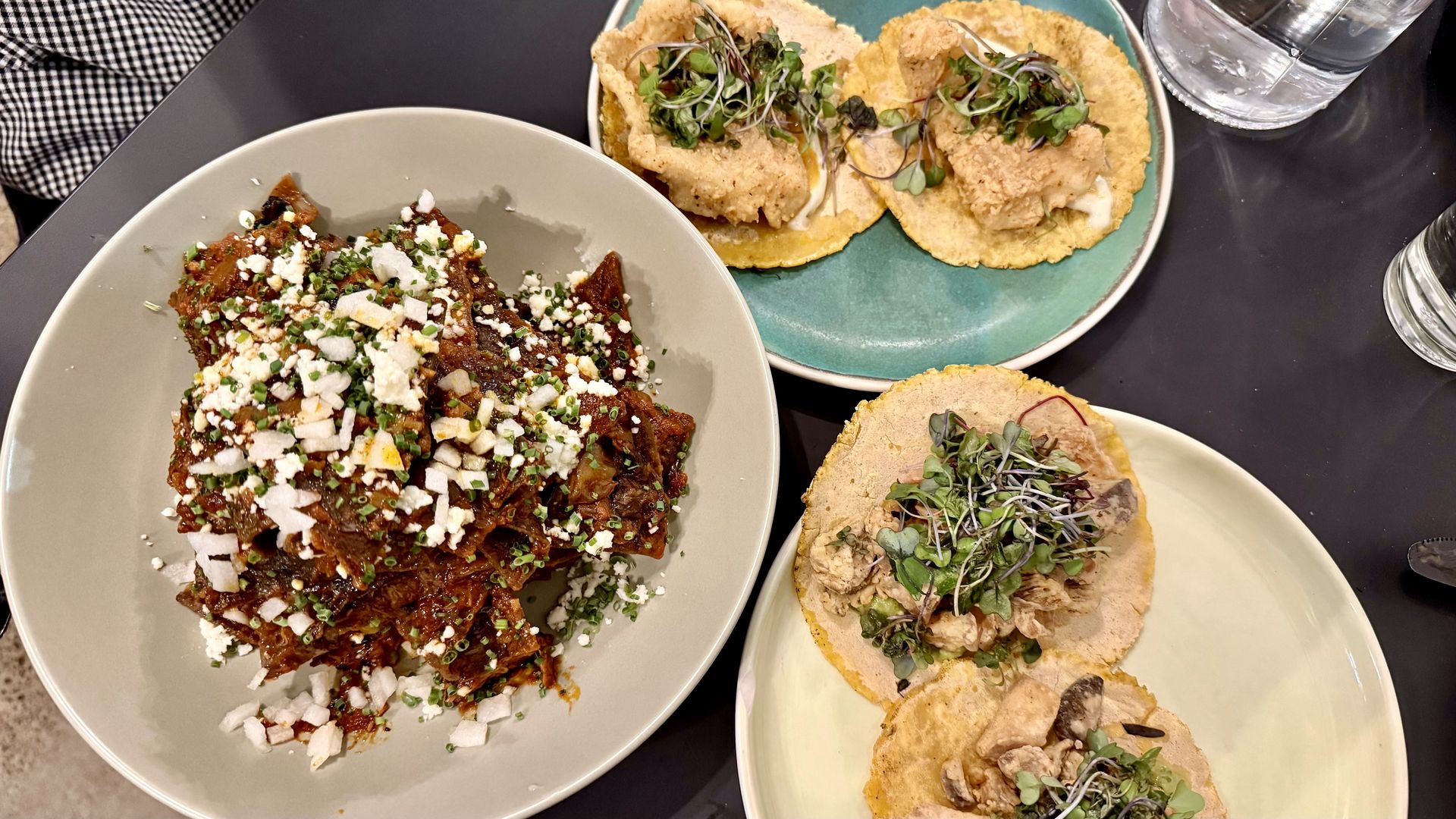 A table with Mexican dishes: a plate of red chilaquiles topped with cheese, onions, and herbs, and two plates of tacos with fried fish and shredded chicken, garnished with microgreens.