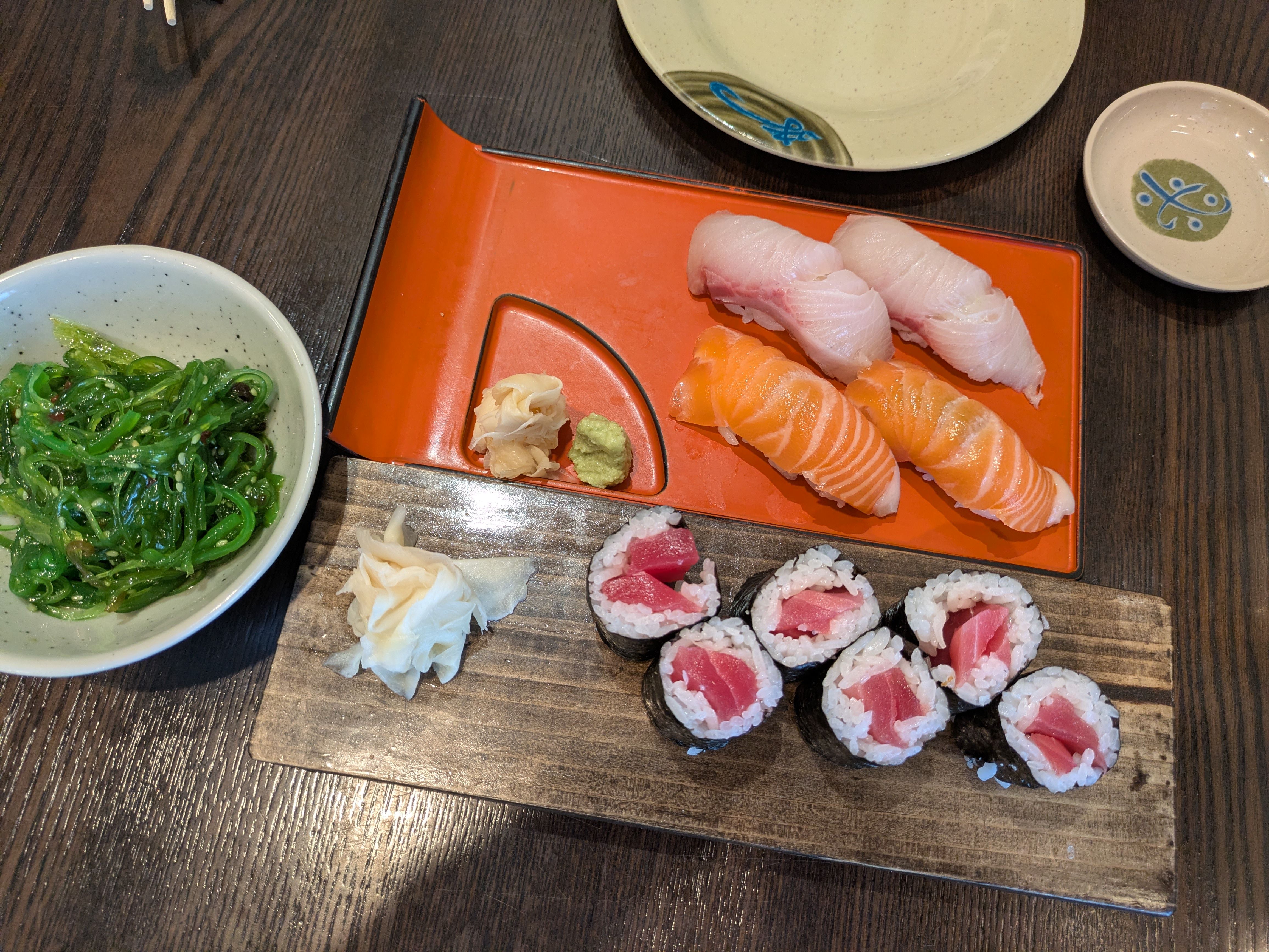 Sushi meal on dark table: salmon and white-fish nigiri on a red tray, tuna maki on a wooden board, and a bowl of seaweed salad with pickled ginger and wasabi.