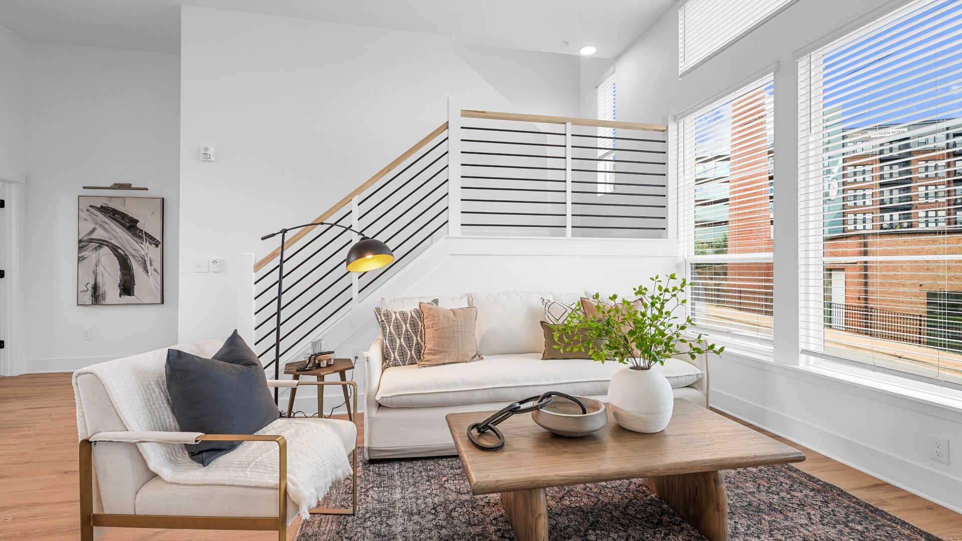 Bright living room with white walls, wood floors, white sofa with mixed beige cushions, armchair with dark pillow, wooden coffee table with green plant, large windows with white blinds.