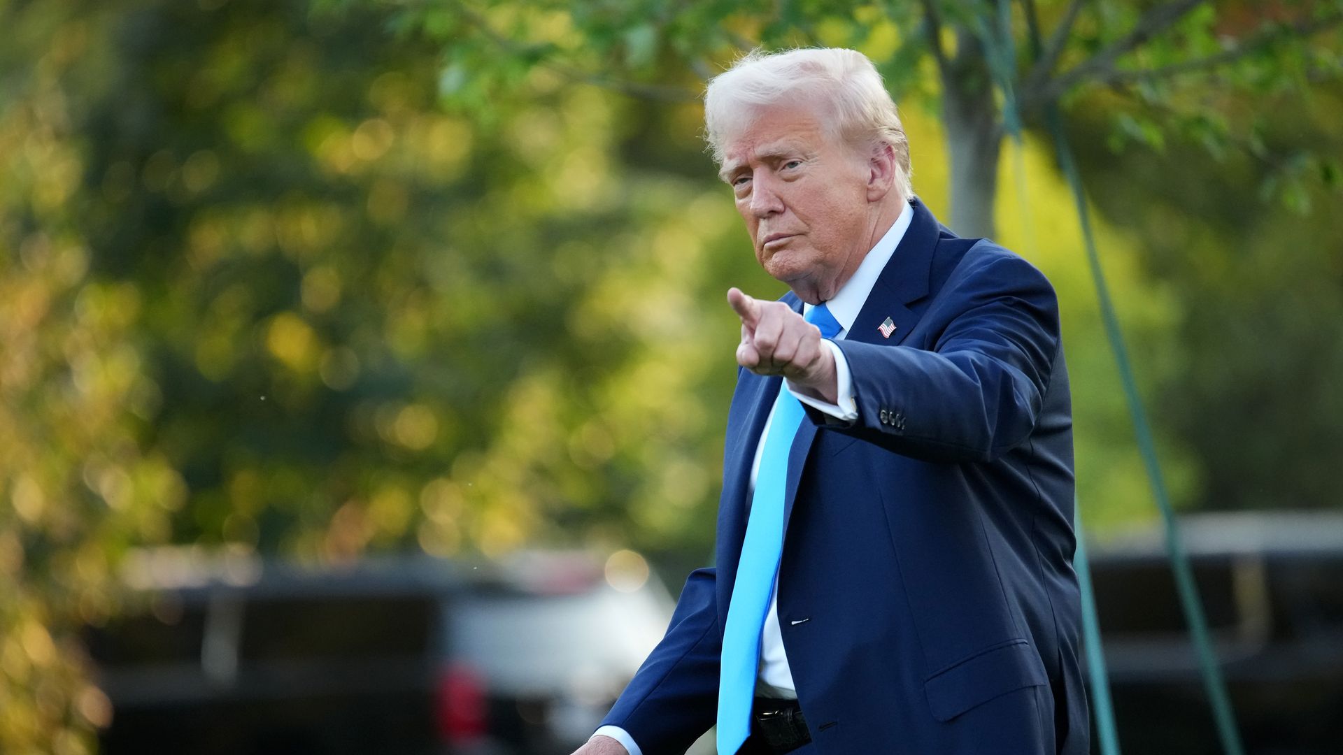 President Trump, wearing a navy jacket with a US flag pin, a white shirt and blue tie, points with his left hand in front of trees.