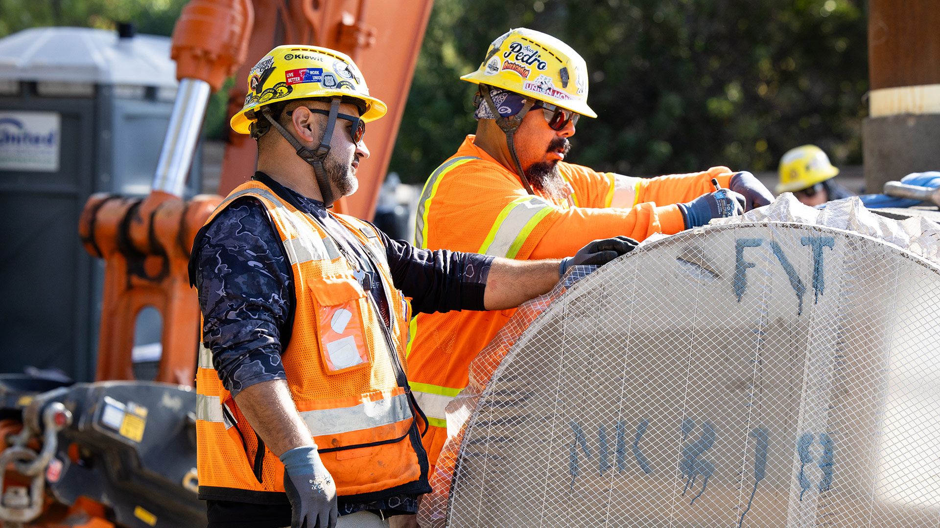 Two construction workers in safety gear and helmets collaborate on a task at an active job site.