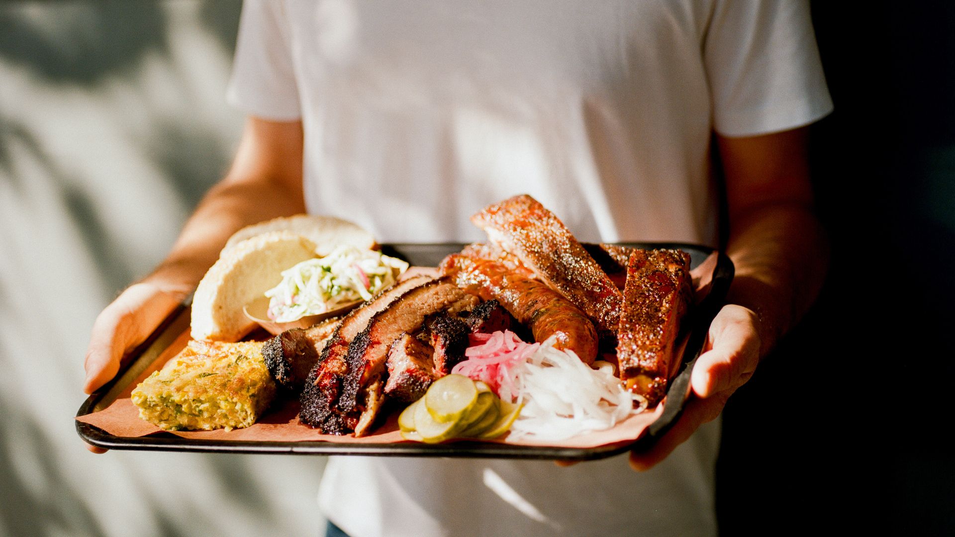 Person in white shirt holding a tray with barbecue ribs, sausage, sliced brisket, pickles, onions, pickled vegetables, cornbread, and a sandwich with coleslaw.