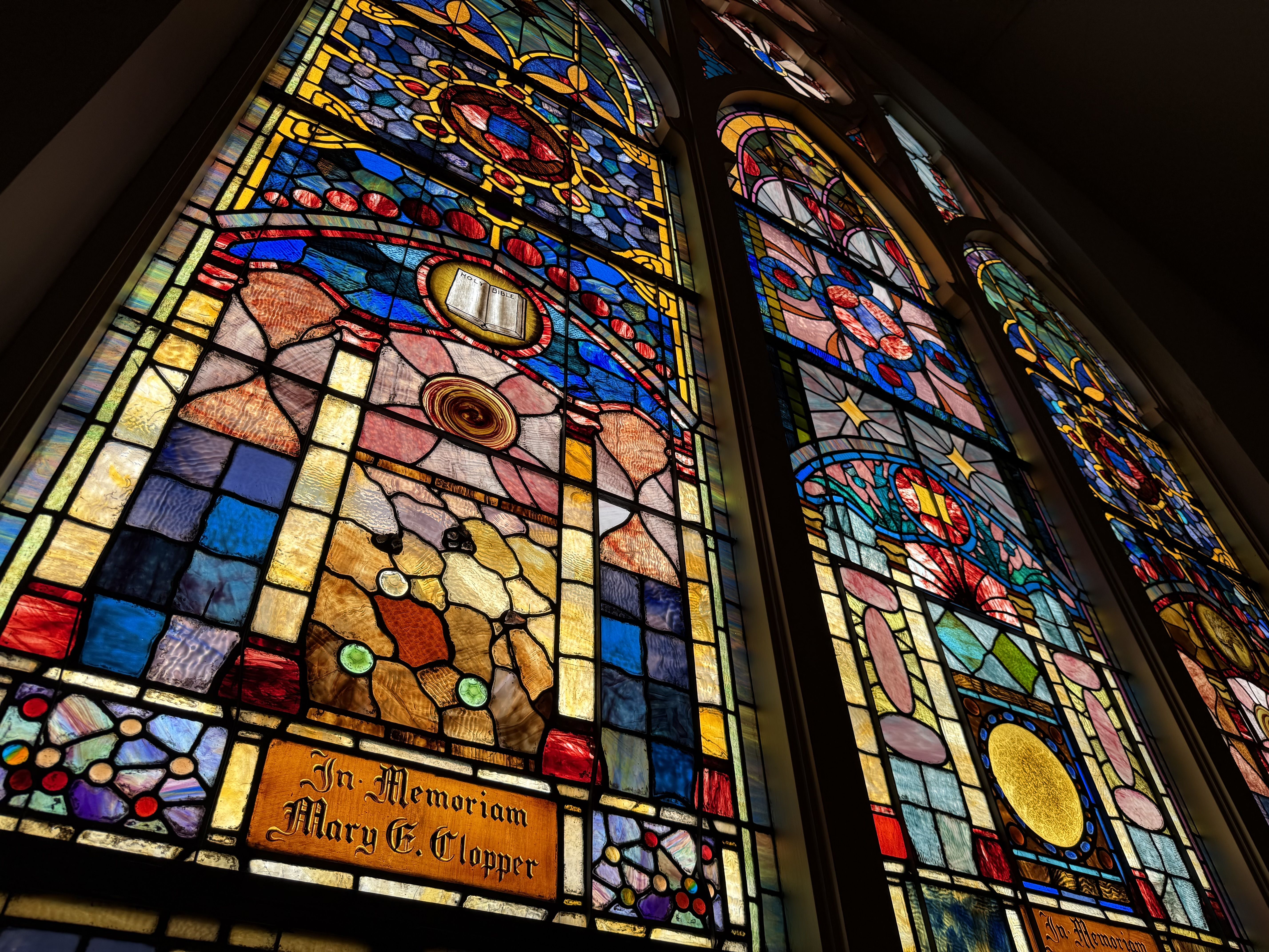 Colorful stained-glass church windows with intricate blue, red, and gold geometric patterns; an 'In Memoriam' plaque with a name visible at the bottom.