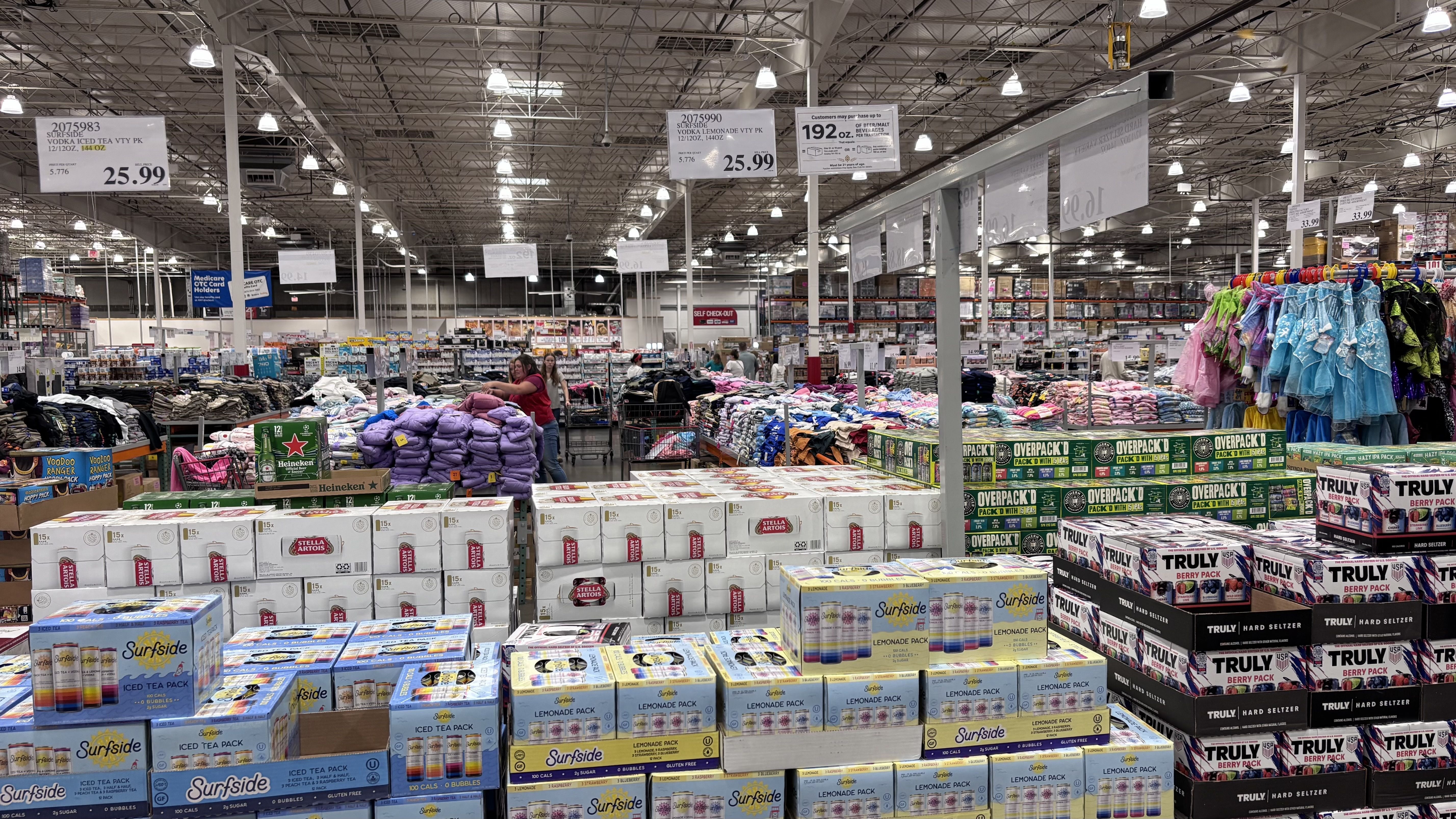 Warehouse-style supermarket interior with bright overhead lights and hanging price signs. Beverages stacked on pallets in the foreground; colorful coats on racks to the right; shoppers browse the aisles.