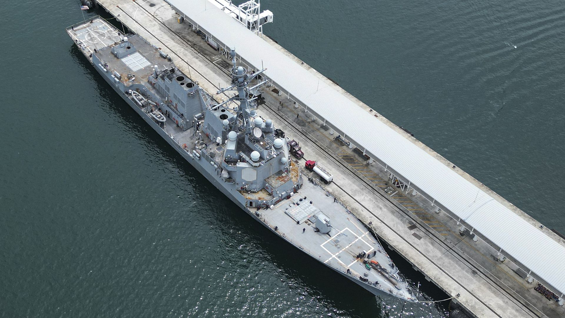An aerial view of a U.S. warship docked. The water is a greyish blue.