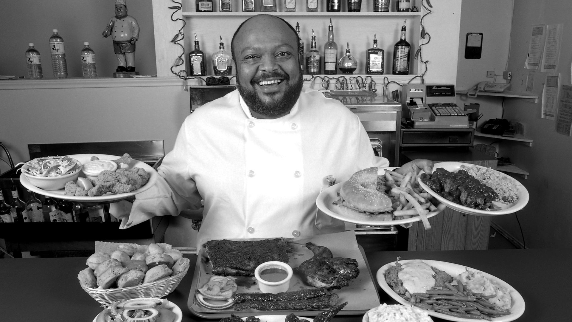 Black-and-white image of a smiling chef in a white coat behind a counter, presenting two plates of fried foods. Shelves with bottles and a cash register are visible in the background.