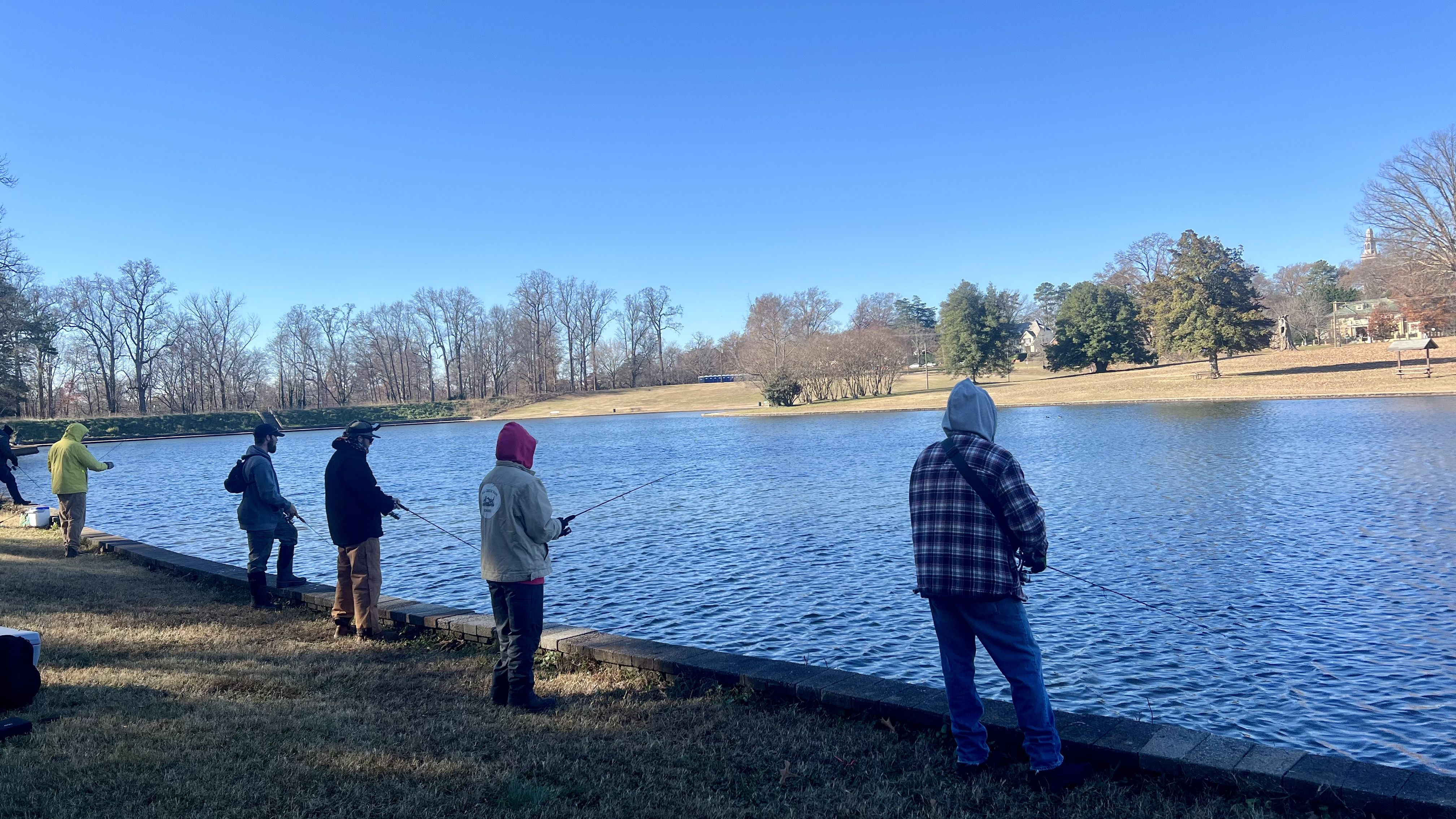 Several people wearing jackets and hoodies fishing along the edge of a calm blue lake on a clear, sunny day with bare trees and green bushes in the background.