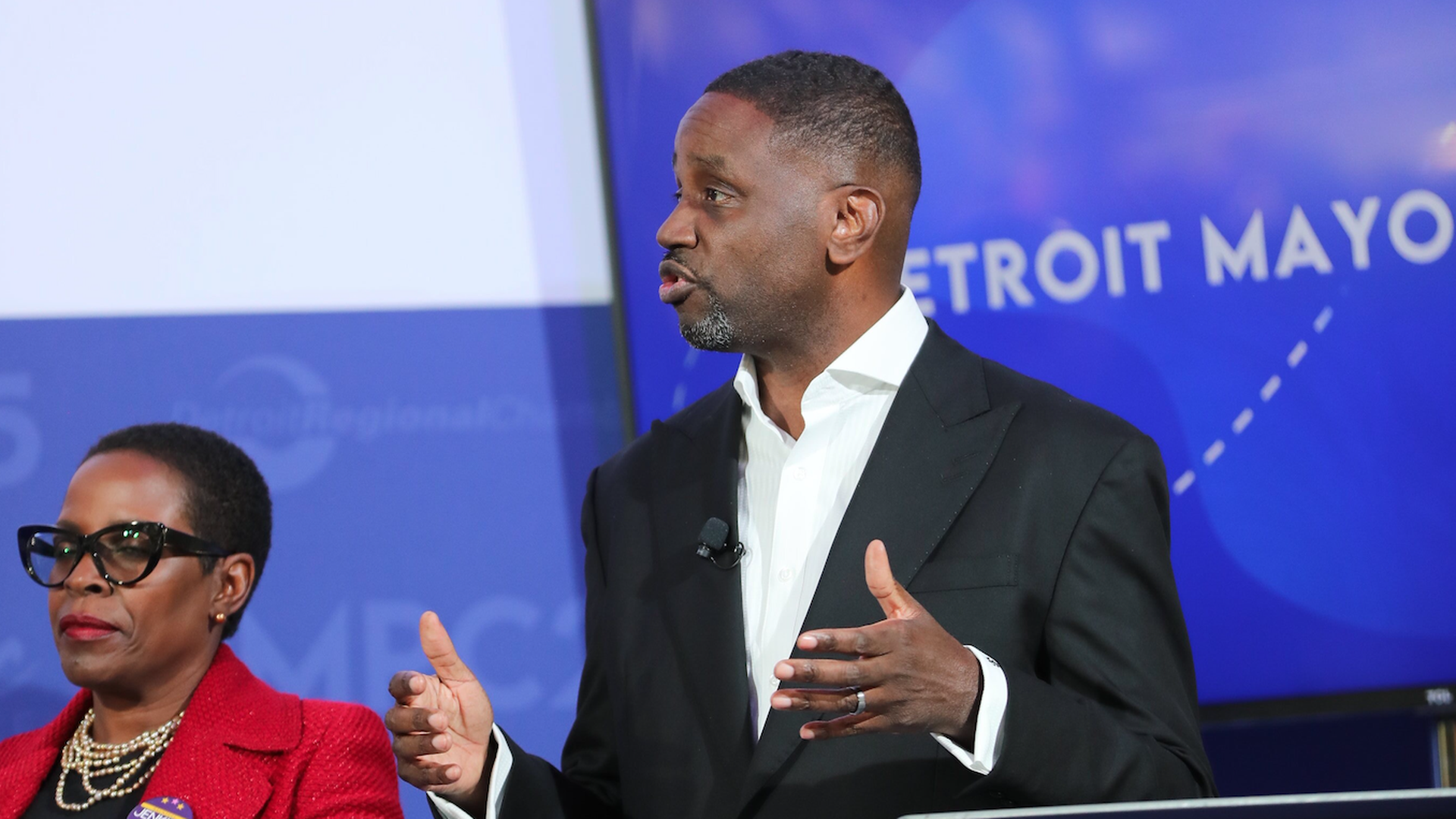 Kinloch at a podium with a blue backdrop, gesturing while wearing a suit