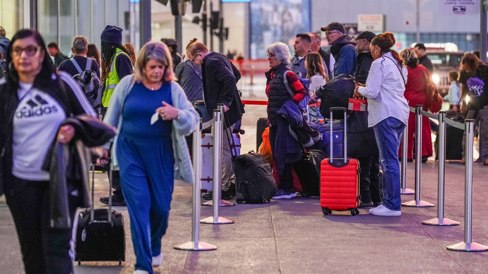 Travelers with wheeled suitcases walk and wait for service at the curbside check-in at an airport. 