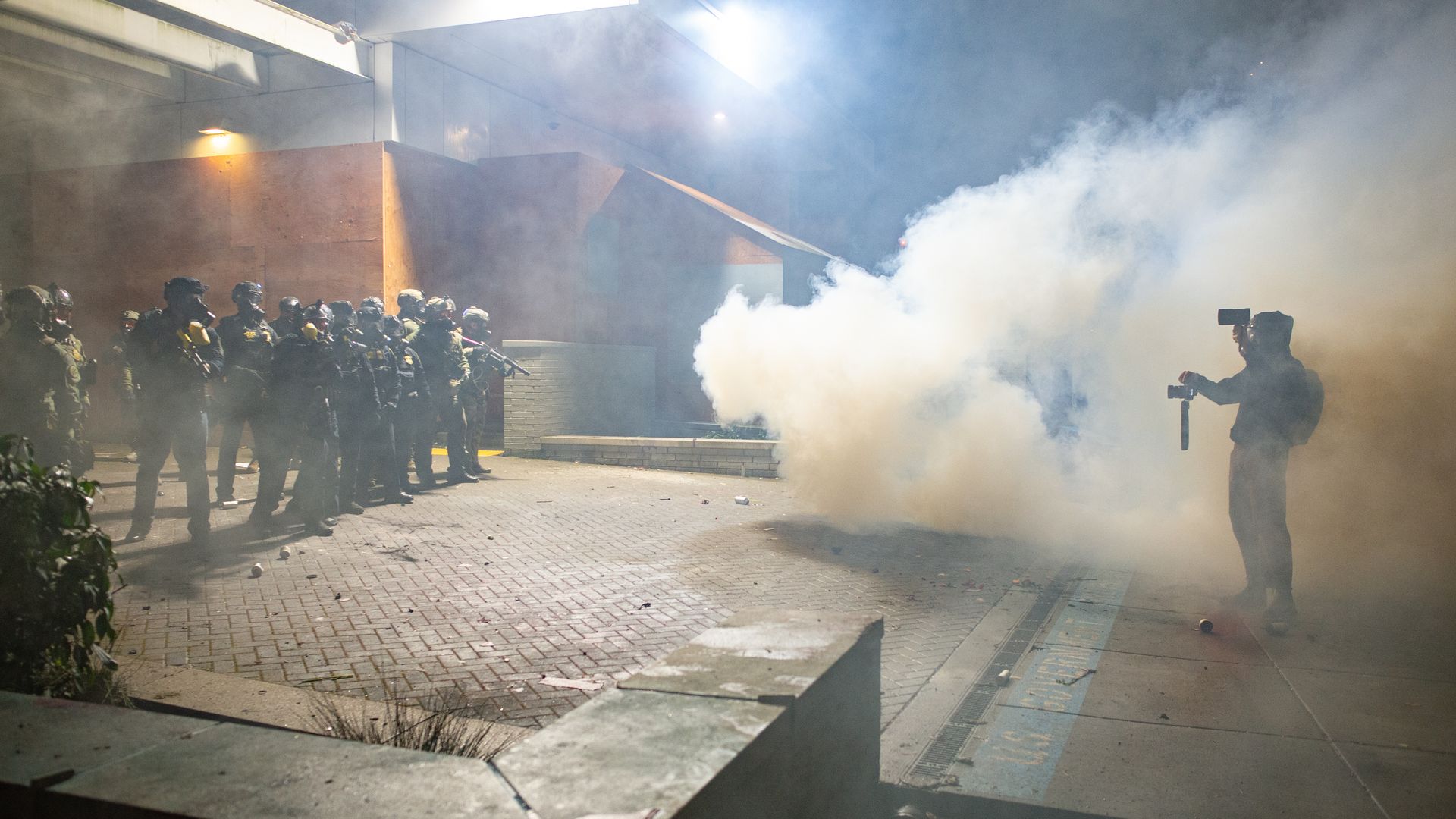 Tense nighttime scene with a line of riot police in helmets and gear facing a single person standing in smoke holding recording devices in a dimly lit urban area.