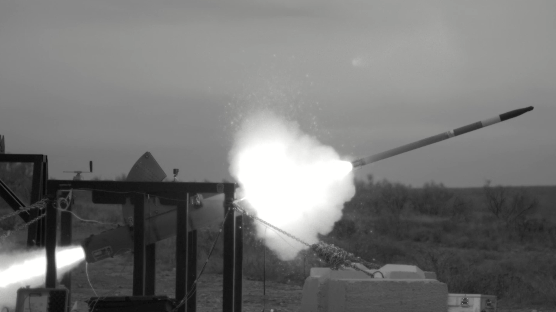 Black-and-white photo of a rocket launch on a test pad, with a bright exhaust plume and a tilted launch rail to the right.