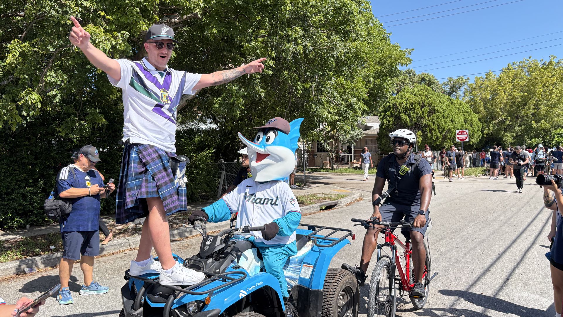 A Scotland suporter wearing a purple tartan kilt stands atop an ATV driven by the Miami Marlins mascot.