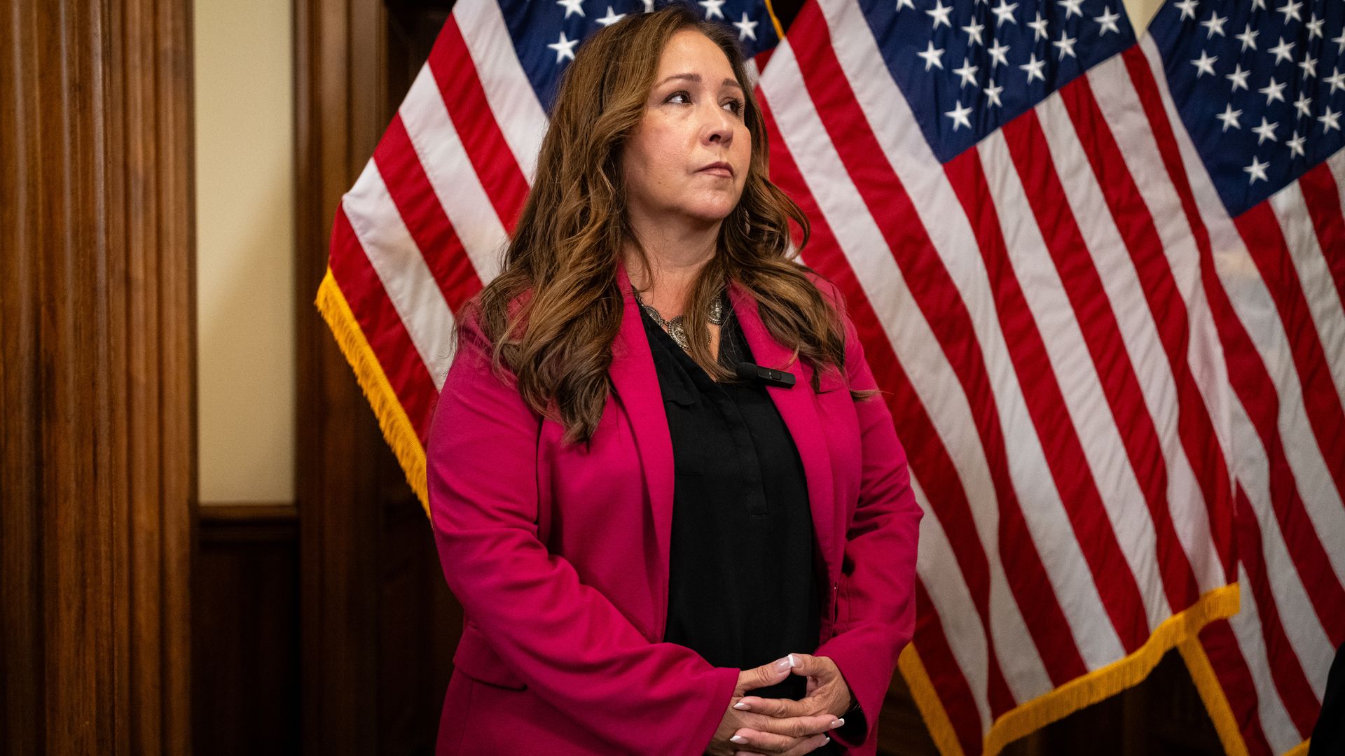 Woman in a magenta blazer and black shirt stands with hands clasped, in front of multiple American flags with wood-paneled walls in the background.