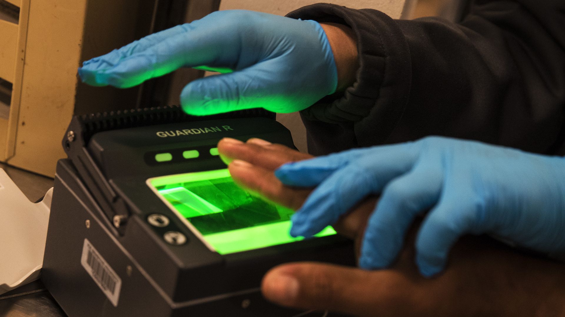 Gloved hands pressing another ungloved hand to a fingerprint scanner