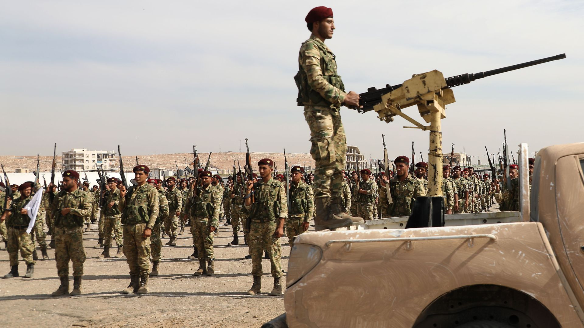 In this image, a soldier stands in the back of a truck while holding onto a stationary machine gun while rows of soldiers stand behind him 