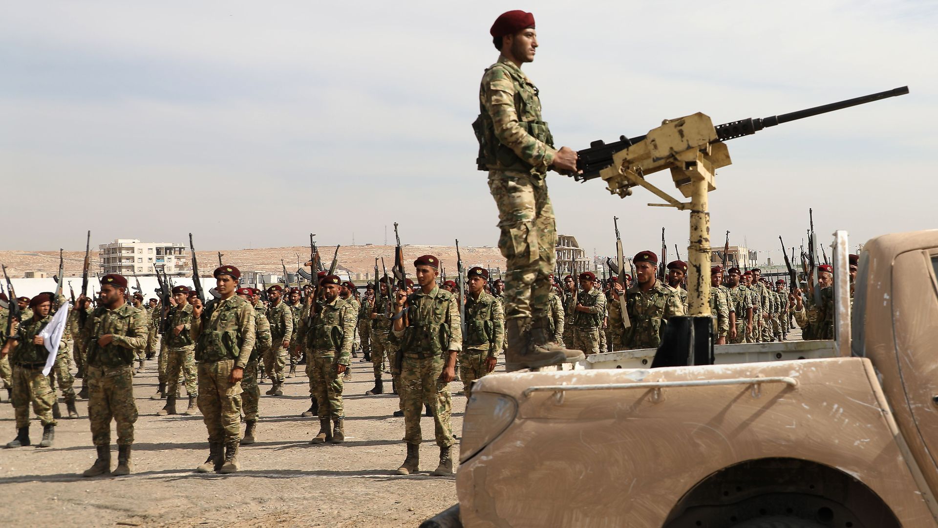 In this image, a soldier stands in the back of a truck while holding onto a stationary machine gun while rows of soldiers stand behind him