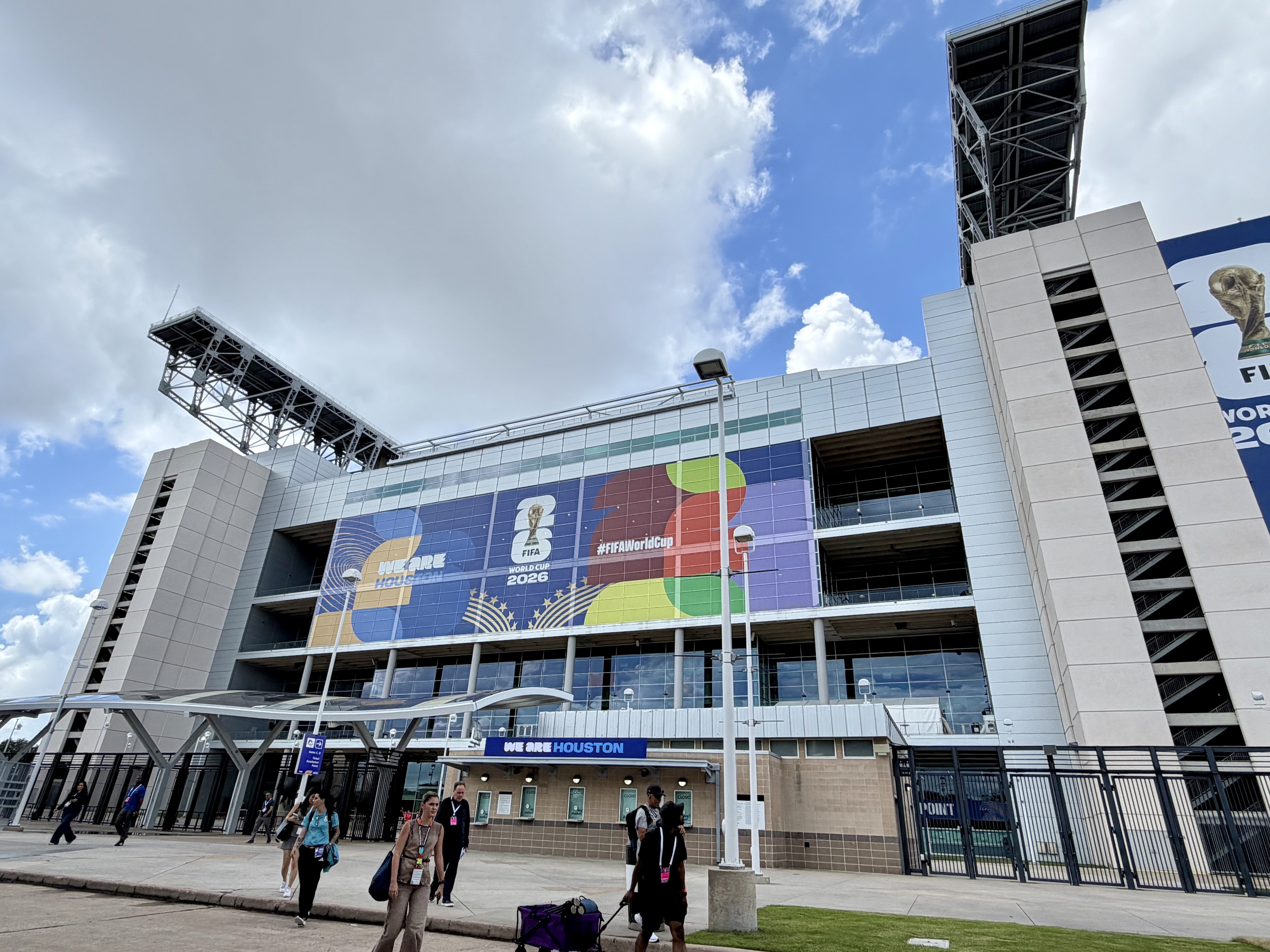 Exterior of a modern stadium with tall lattice towers and colorful banners for the FIFA World Cup 2026; people walk along the foreground under a partly cloudy blue sky.