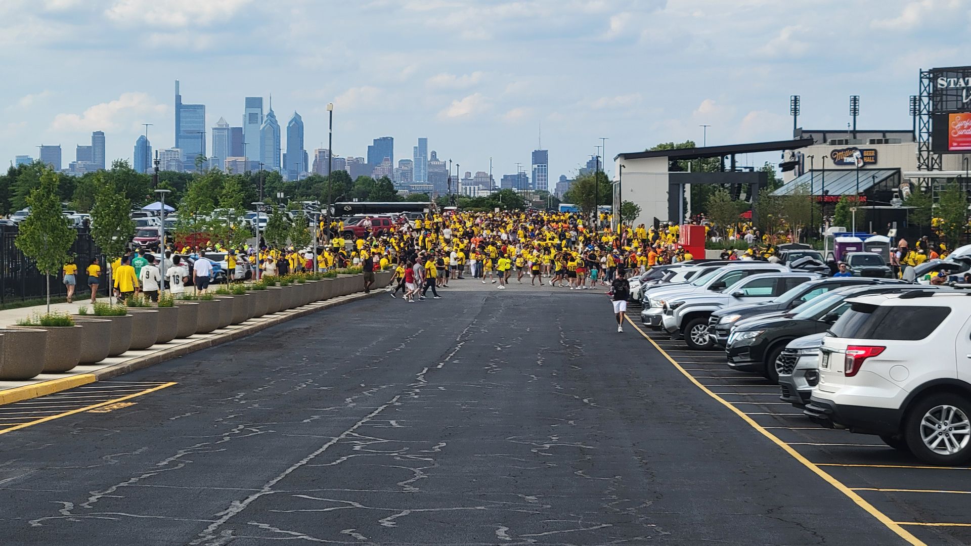 Large crowd in yellow shirts gathers in a parking lot near Philadelphia Stadium with a distant city skyline.