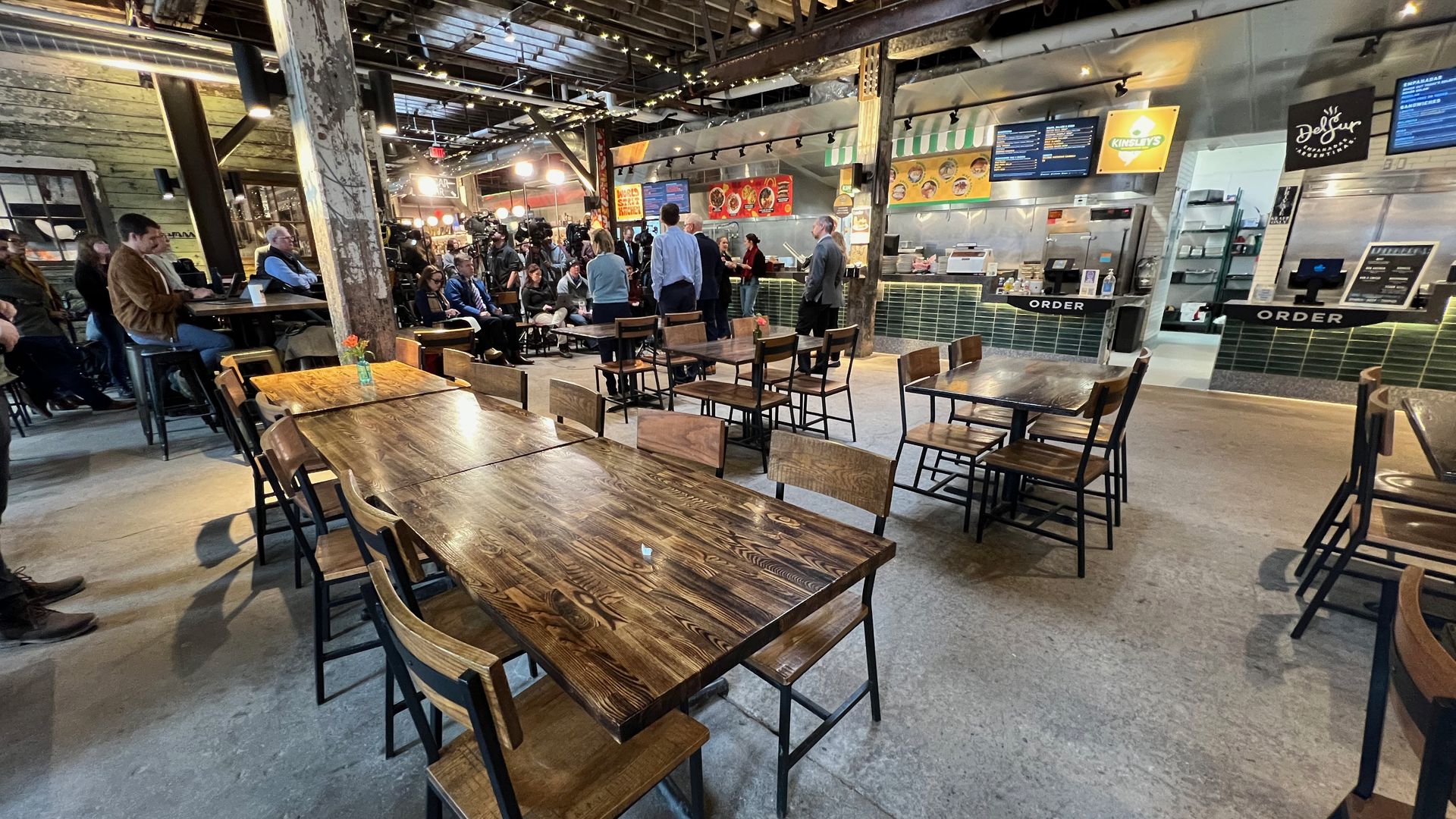 Rustic indoor food hall with long wooden tables and chairs, string lights, and exposed beams. People gather near the order counters with menu boards in the back.