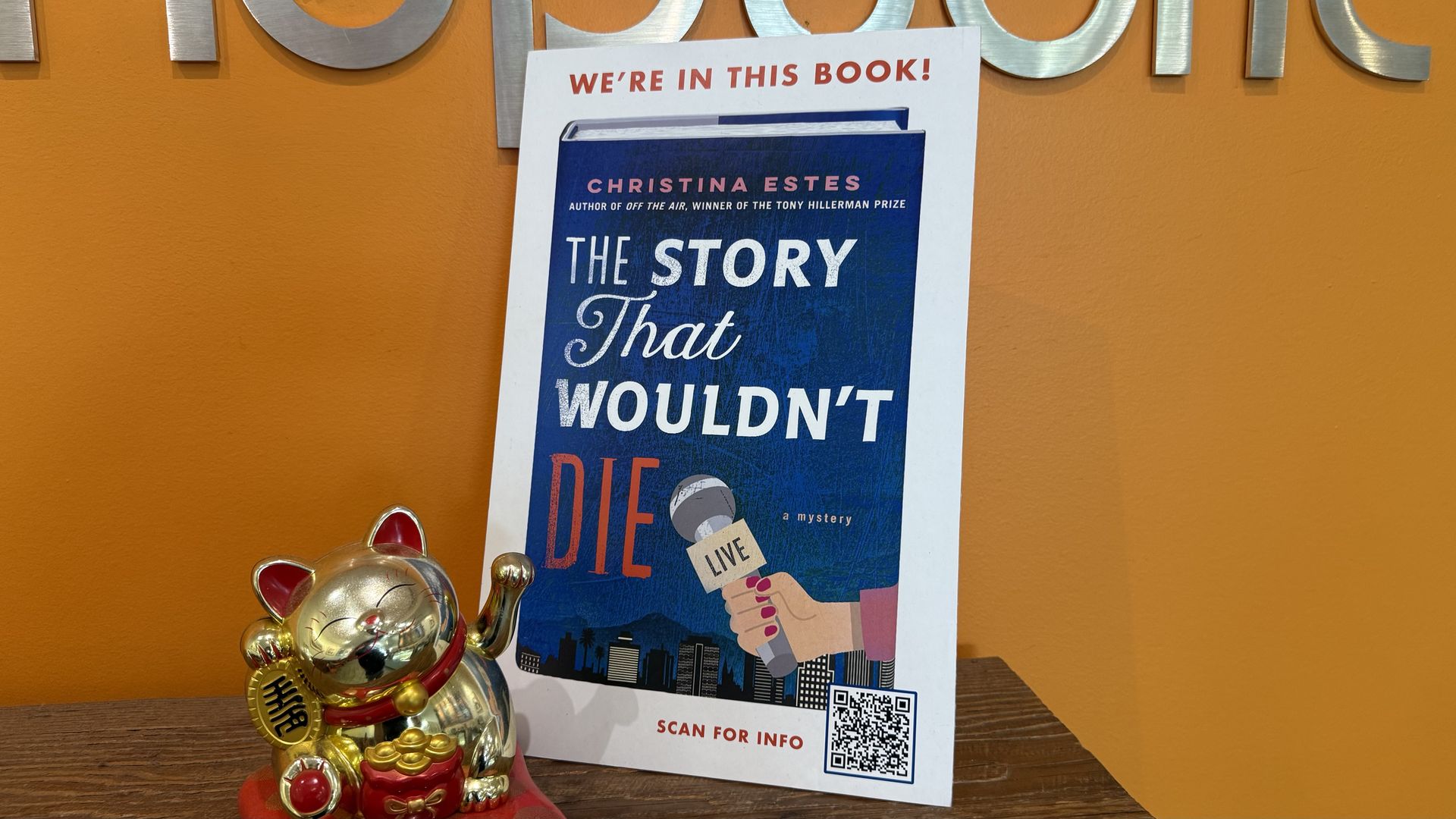 A promotional sign with a blue book cover titled "The Story That Wouldn't Die" by Christina Estes, held by a hand with red nails holding a microphone, under the words, "We're in this book!". A gold lucky cat figurine sits on a wooden surface beside it.