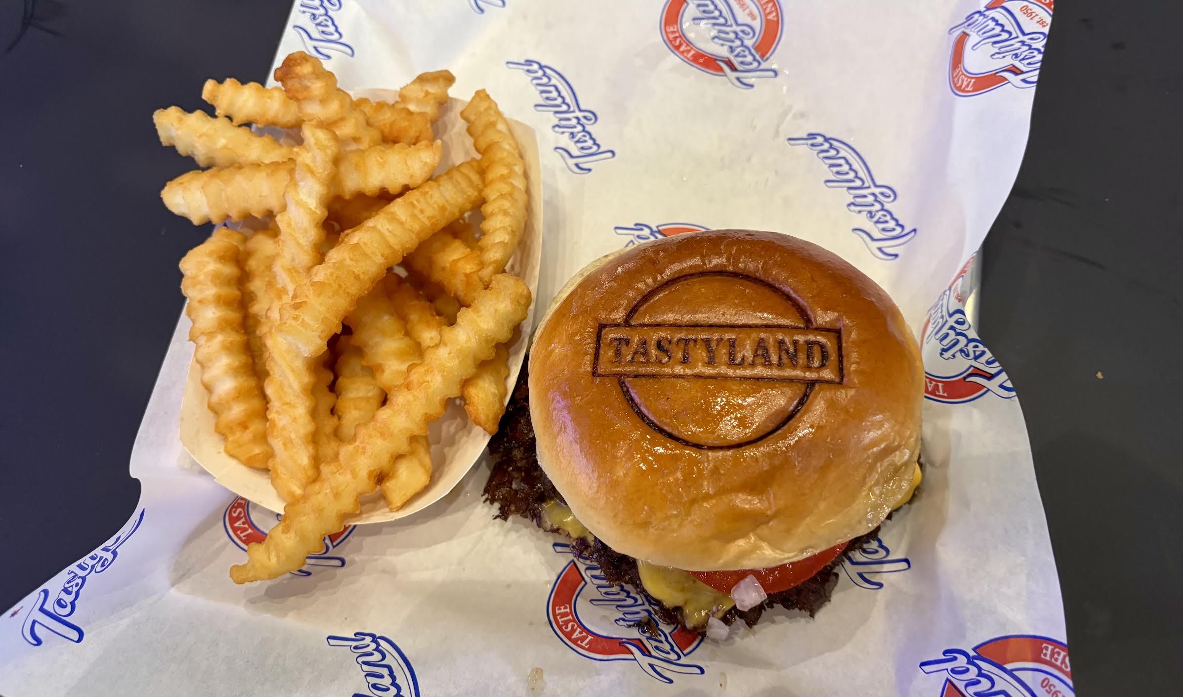 Top-down view of a fast-food meal: crinkle-cut fries in a paper boat beside a glossy burger with the bun stamped "TASTYLAND" on branded parchment.