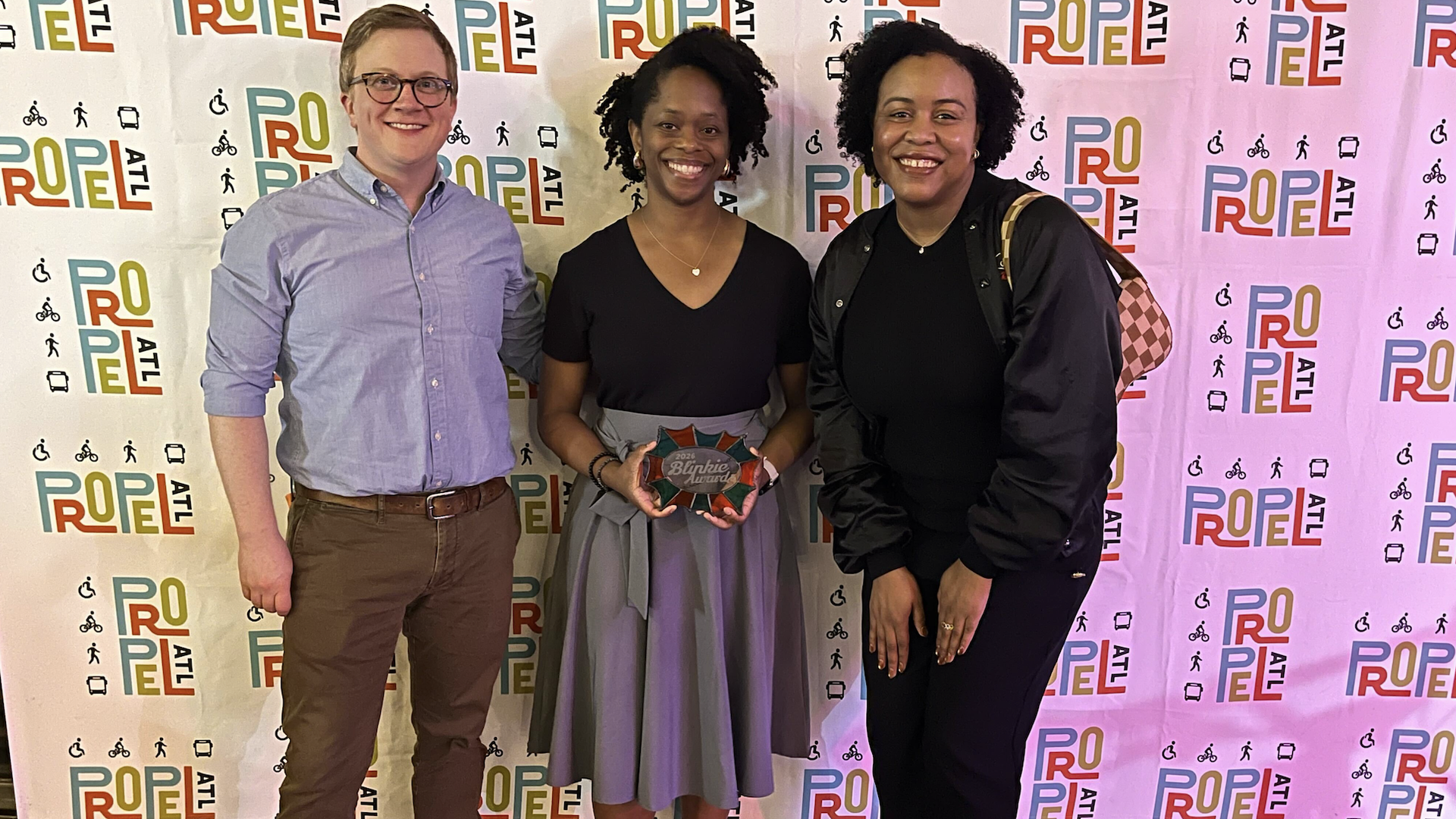 Three smiling people pose before a backdrop of colorful PROPEL ATL logos and accessibility icons. The center person holds a starburst award plaque.