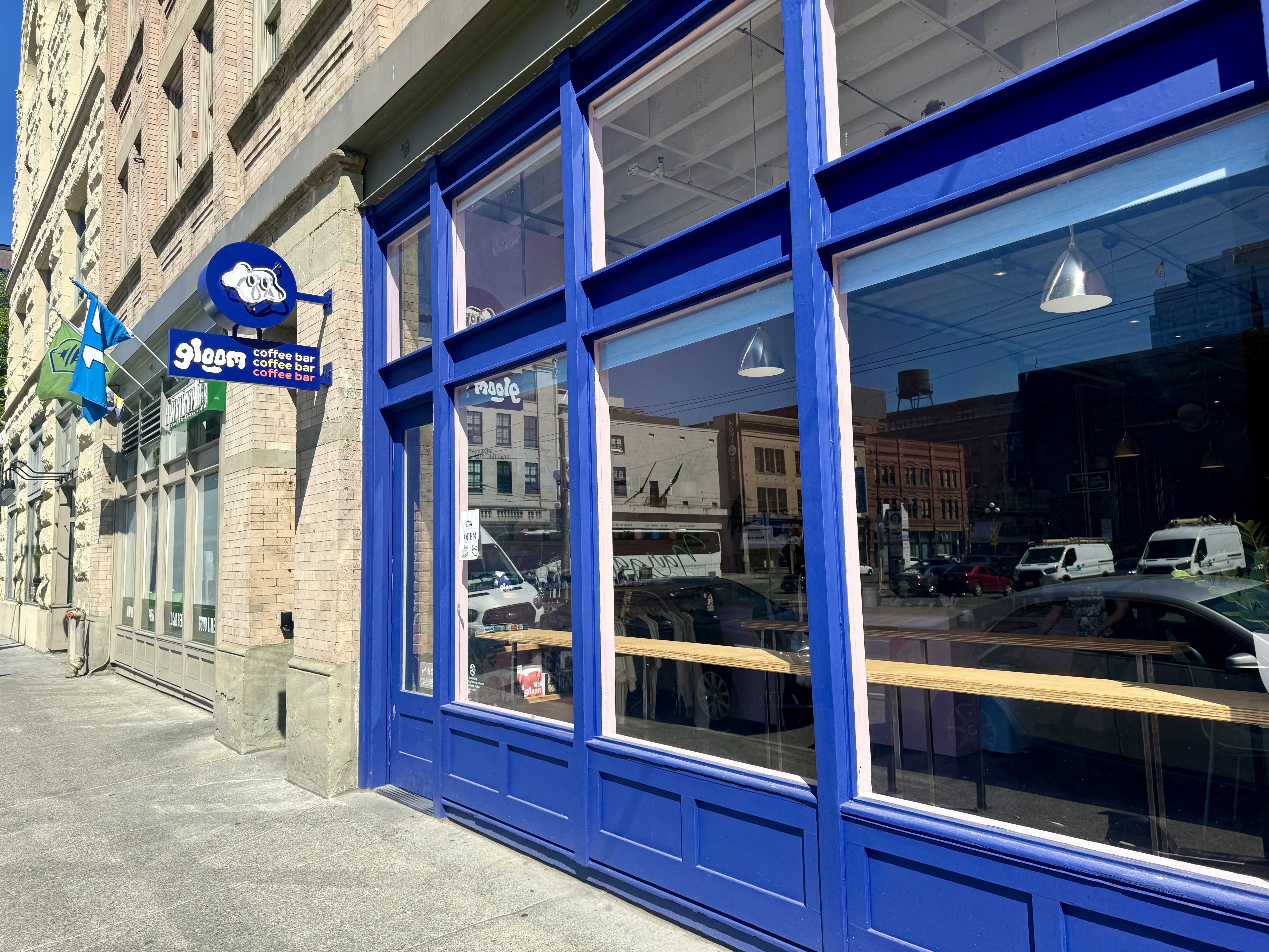 Street-level view of a city block featuring a bright blue storefront with large glass windows and blue frames, a circular blue sign with a white graphic, flags outside, and a coffee bar sign.