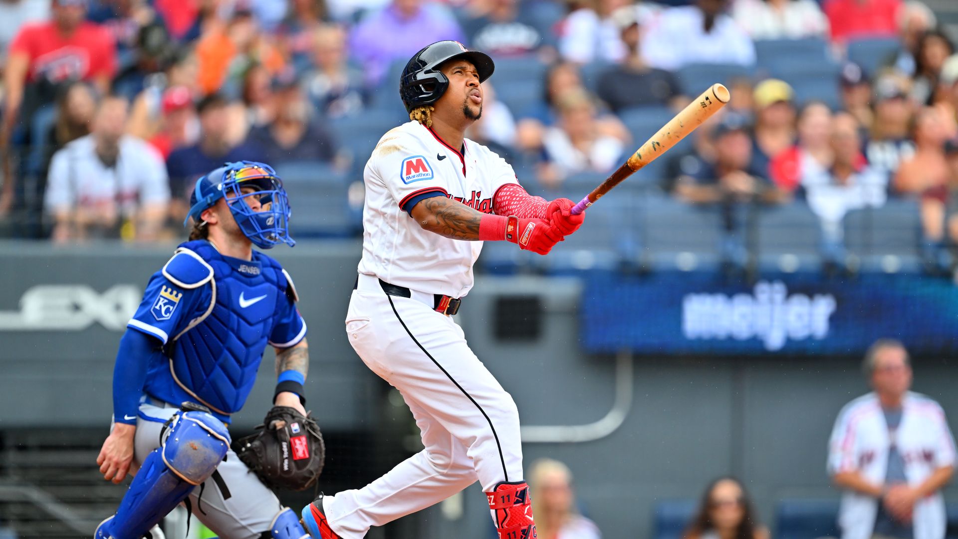 Baseball batter in a white uniform with red trim swings a light brown wooden bat as a blue-gear catcher crouches behind; a blurred crowd watches from the stands.